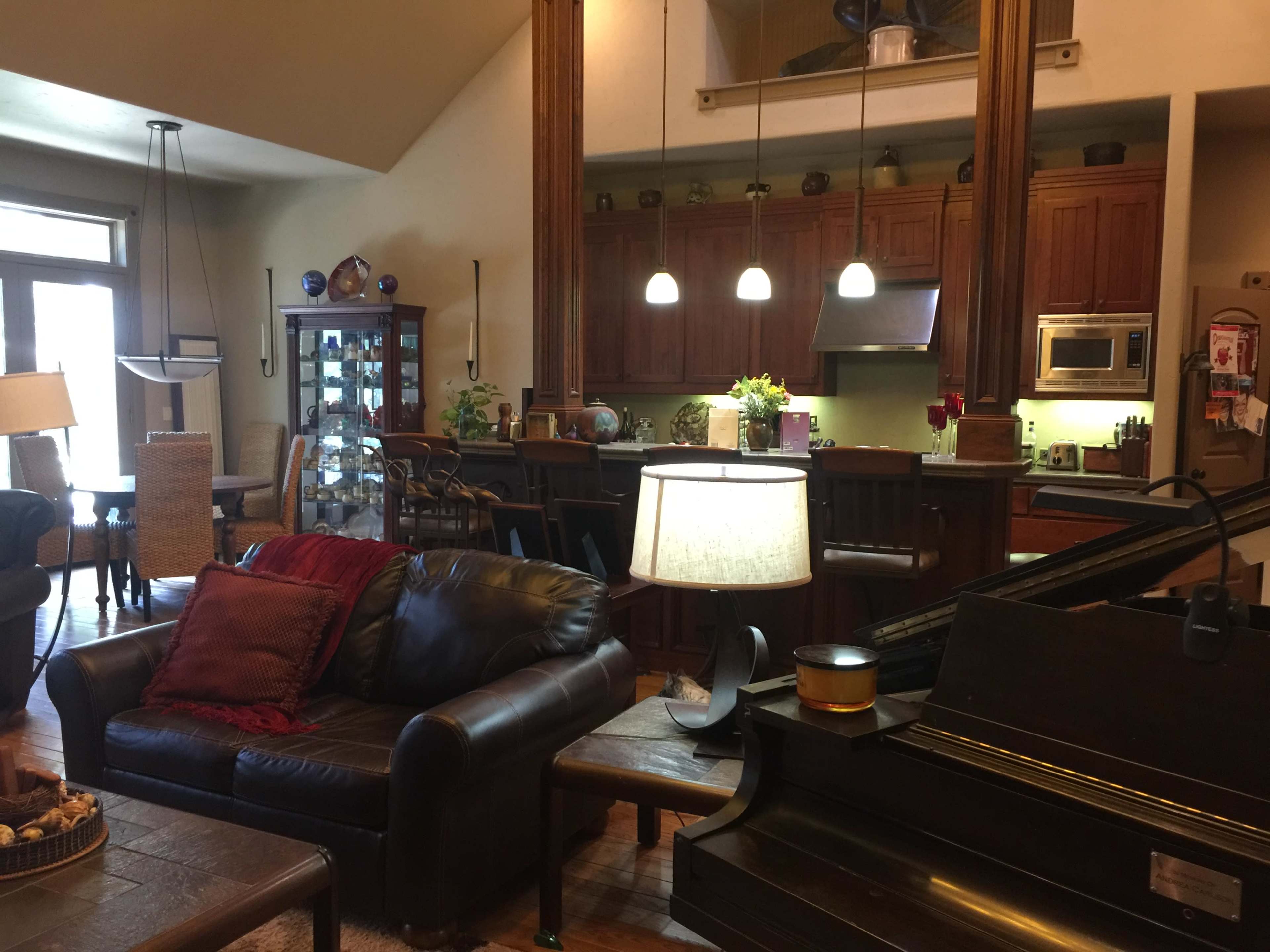 A spacious living room features a leather sofa, a grand piano, and a kitchen area with wooden cabinets and pendant lighting.