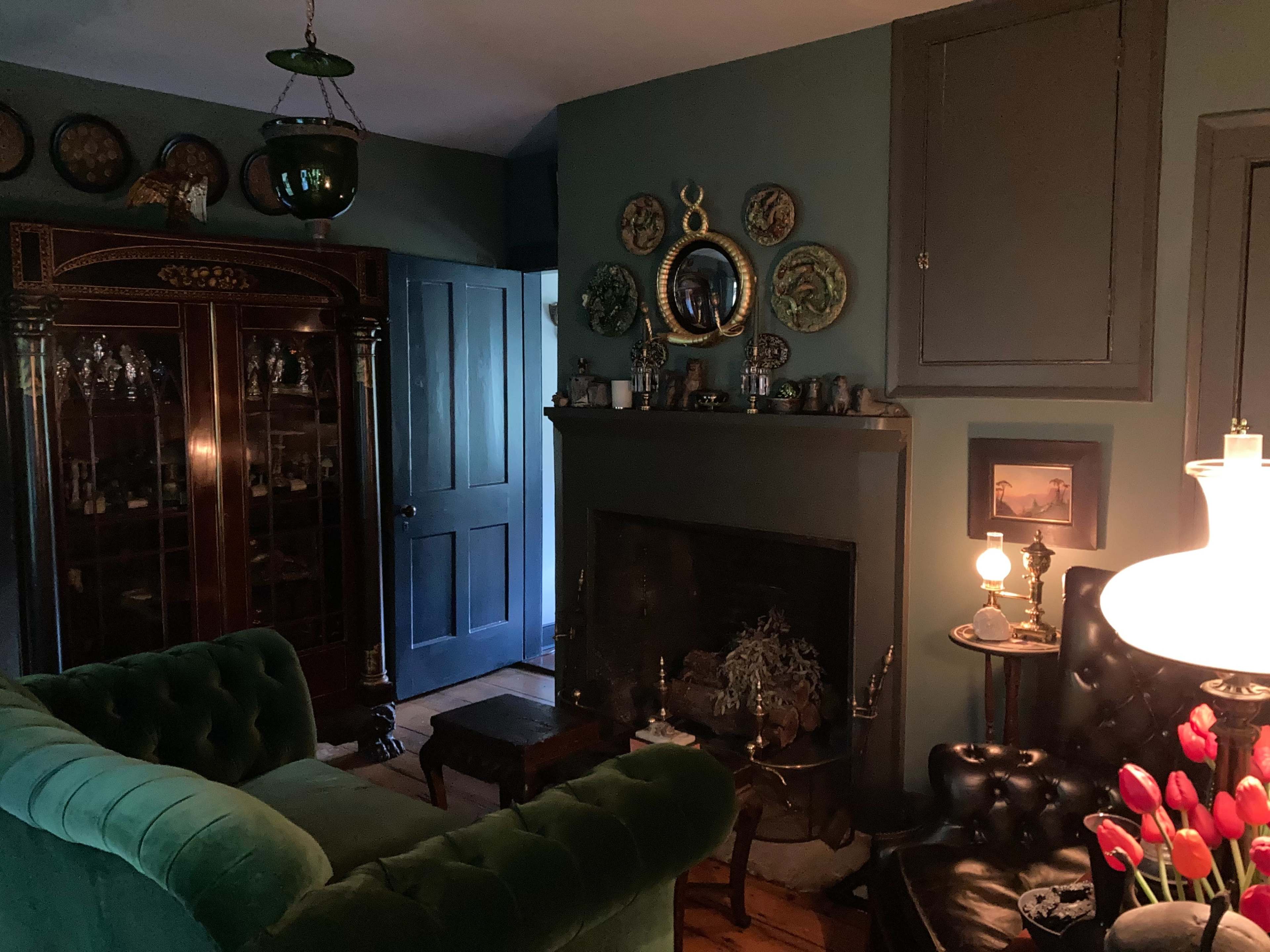 The room features a dark green color scheme with a vintage cabinet, a fireplace adorned with various items, and plush seating arranged around a small table.