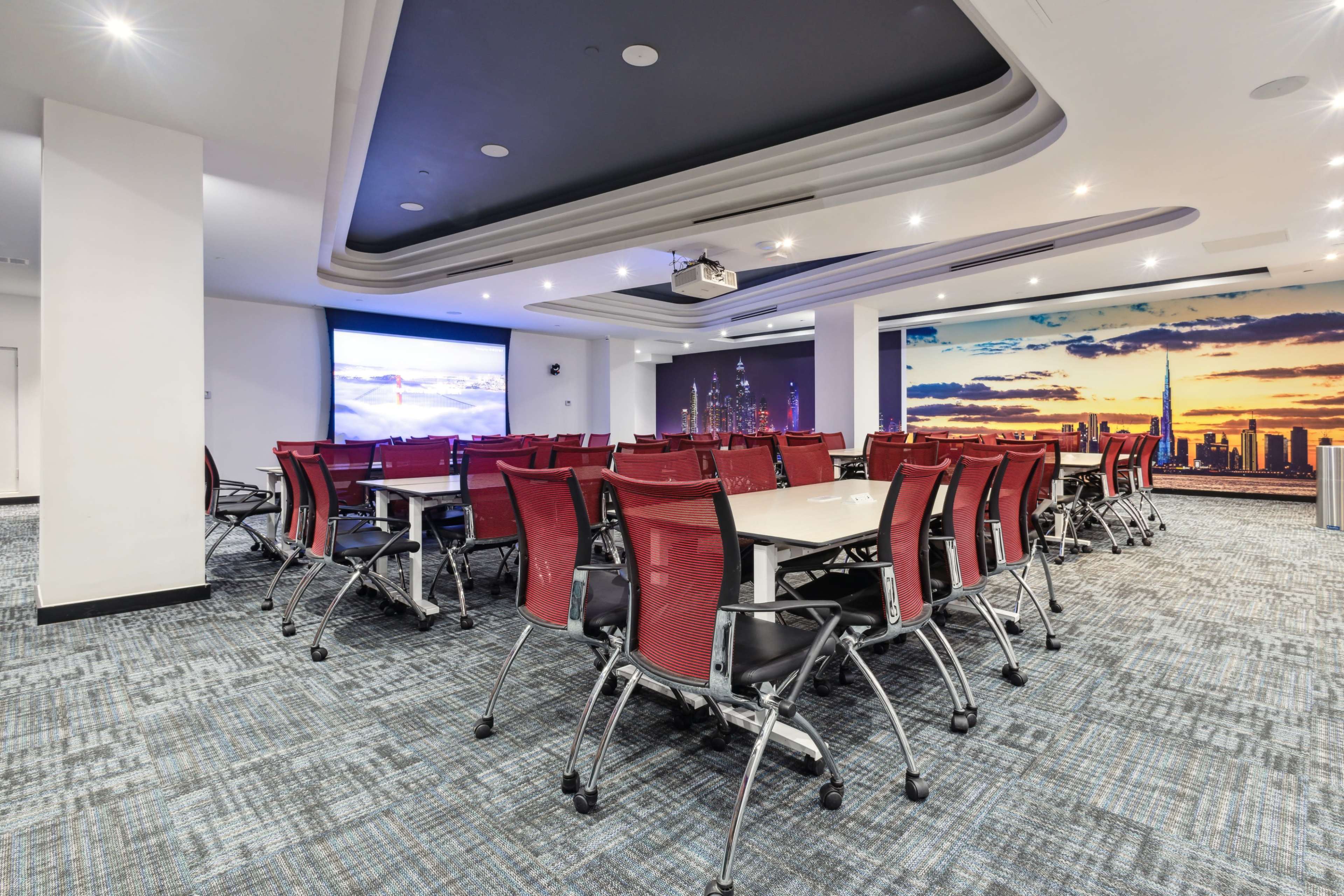 A conference room features rows of red chairs around tables, with large screens displaying cityscape images on the walls.