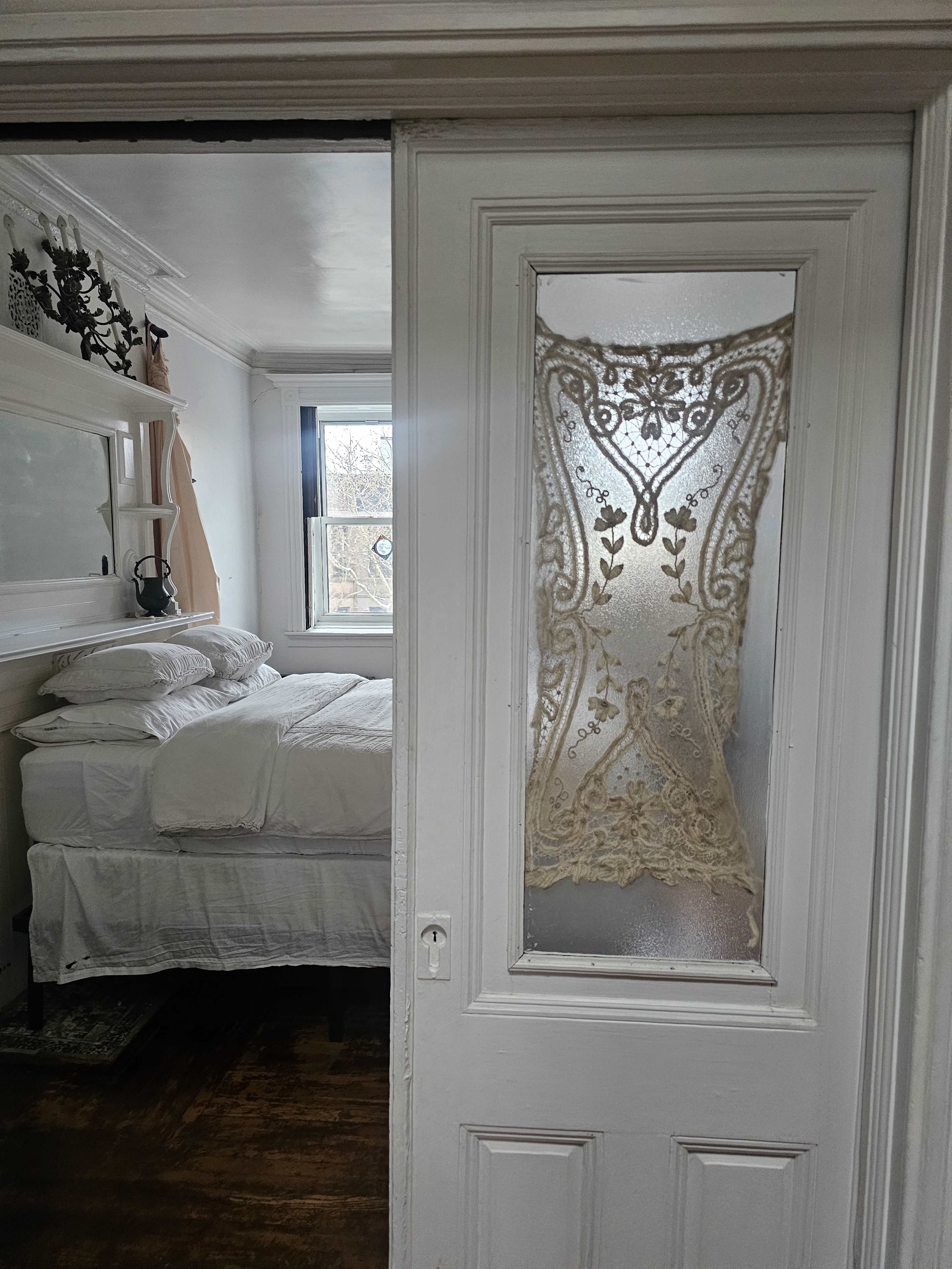 A partially open door reveals a bedroom with a bed covered in white linens and a window in the background.