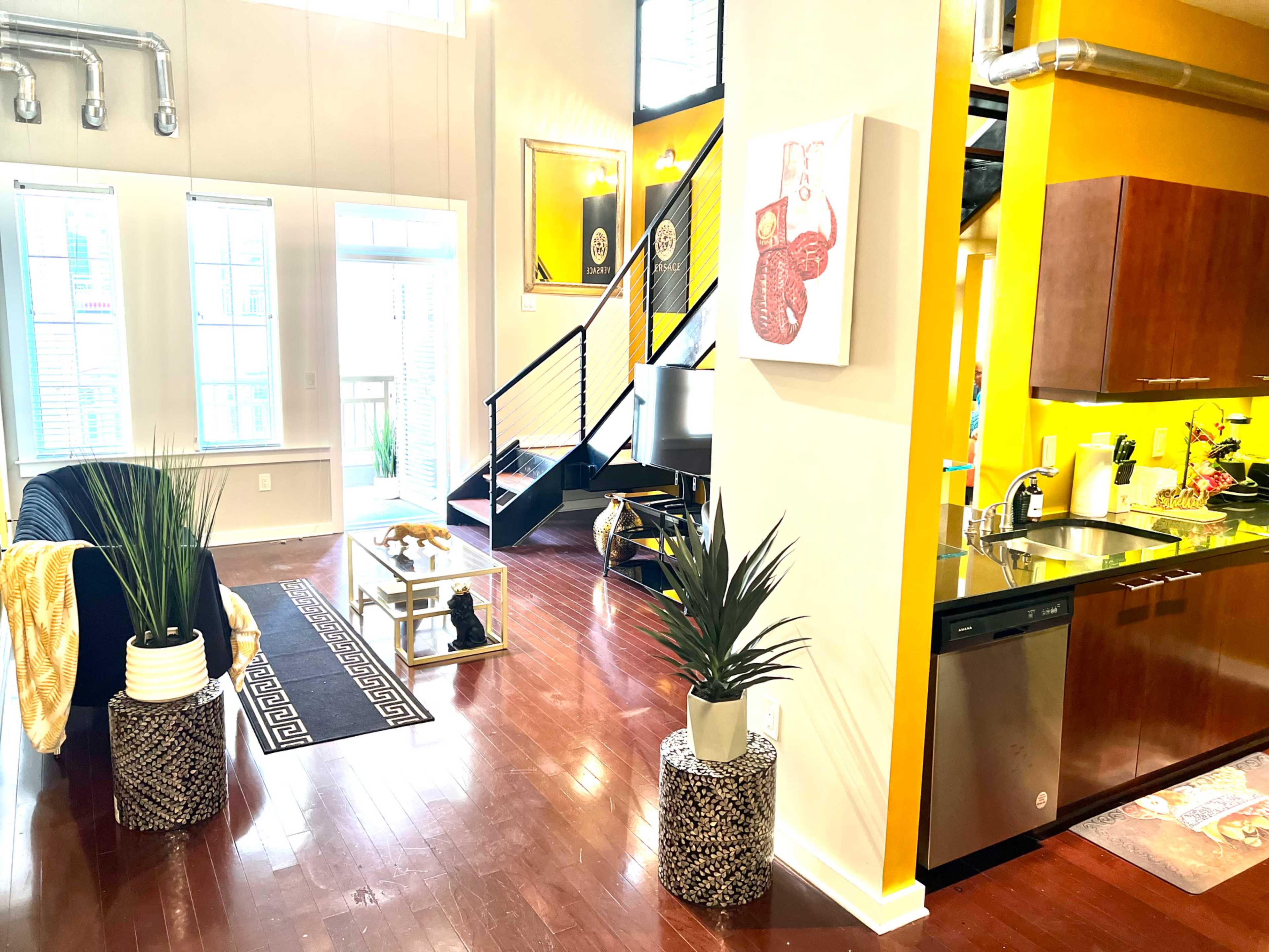 A modern apartment interior featuring a living area with a black couch, a glass coffee table, and kitchen with yellow walls and stainless steel appliances.