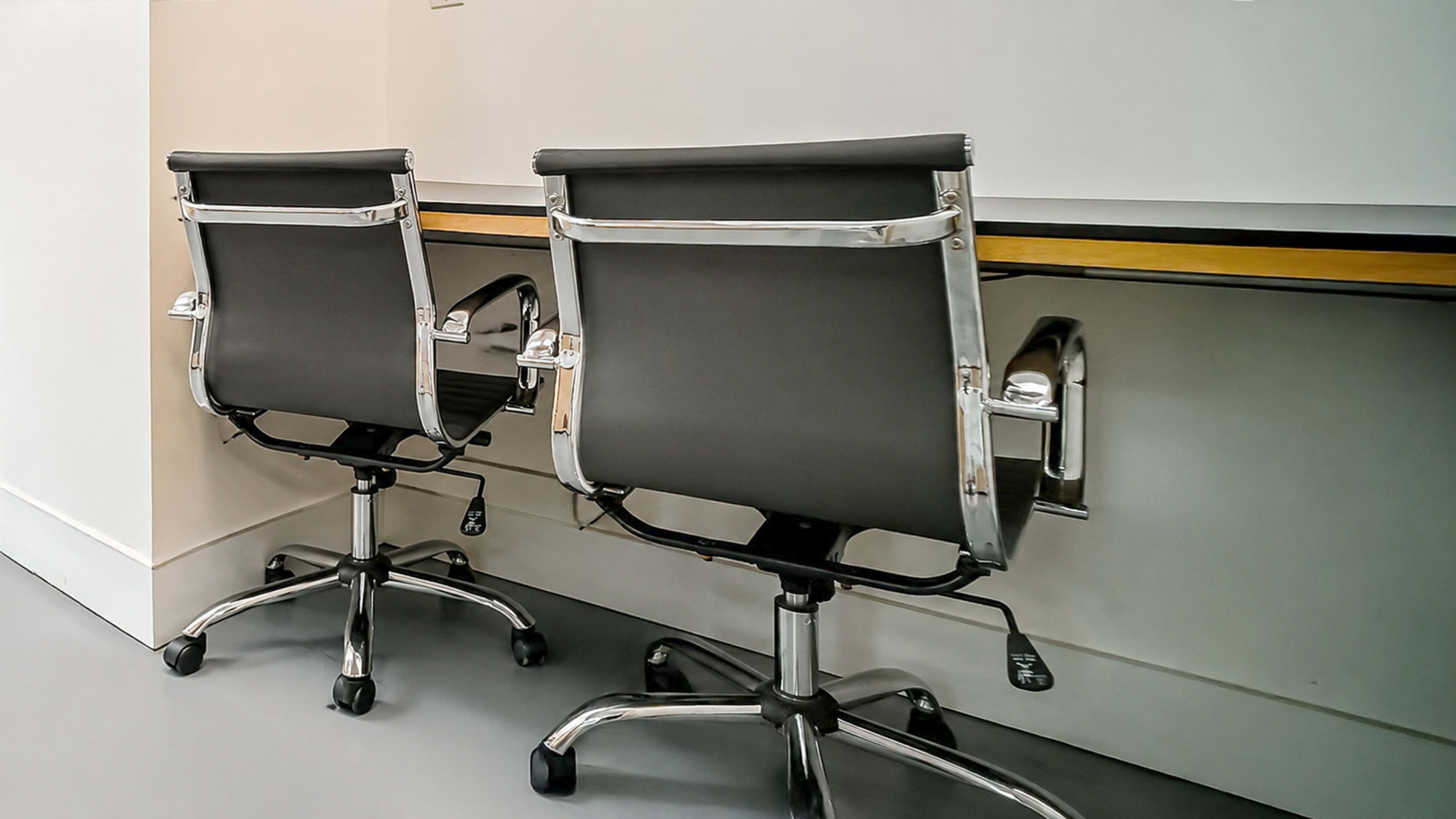 Two black office chairs with chrome frames are positioned against a plain white wall under a long wooden desk.