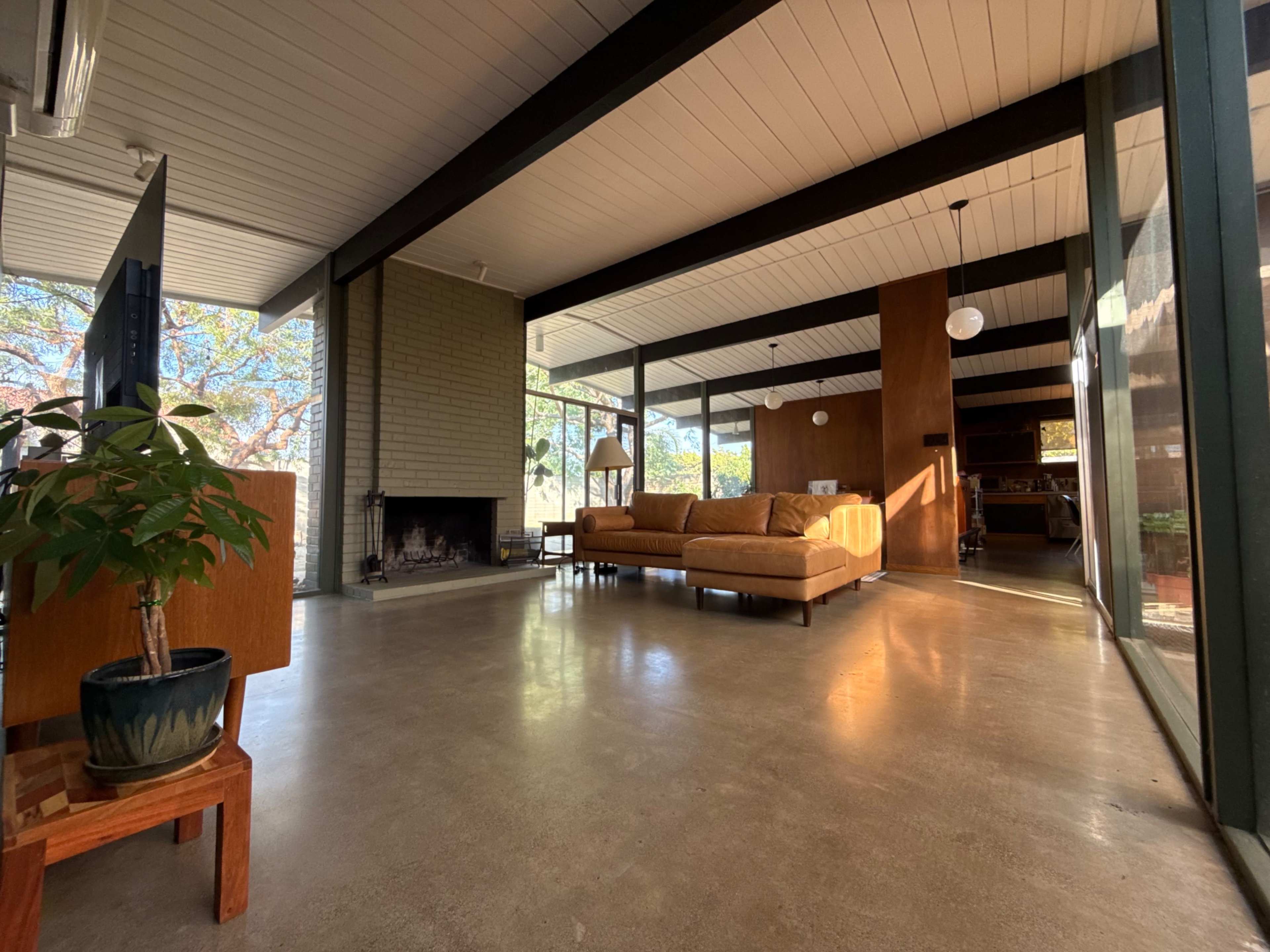 The image shows a modern living room featuring a brown sofa, a fireplace, and large glass windows that provide a view of the outdoors.
