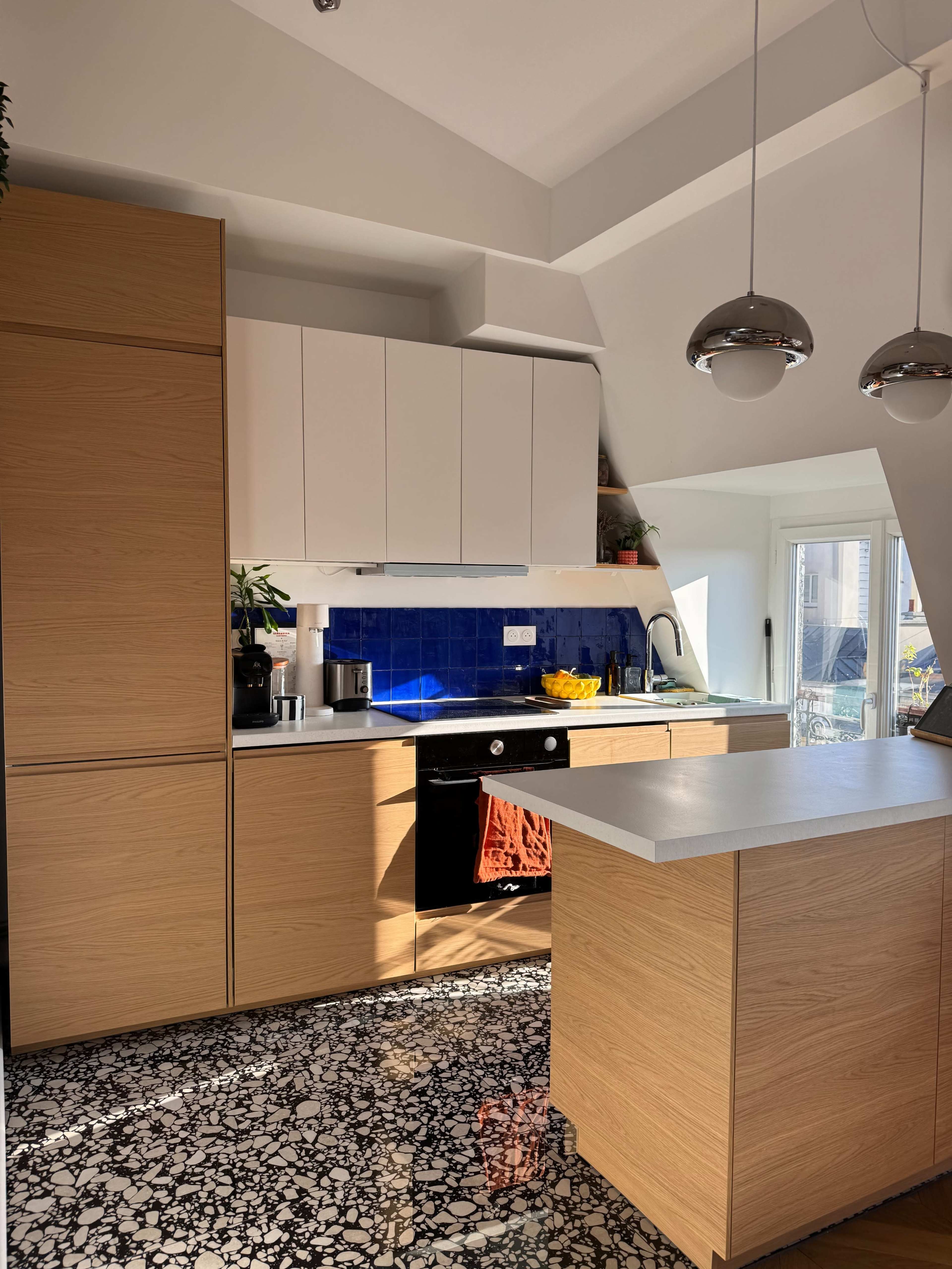 The image shows a modern kitchen with wooden cabinets, a blue tiled backsplash, and a white countertop, featuring a window that lets in natural light.