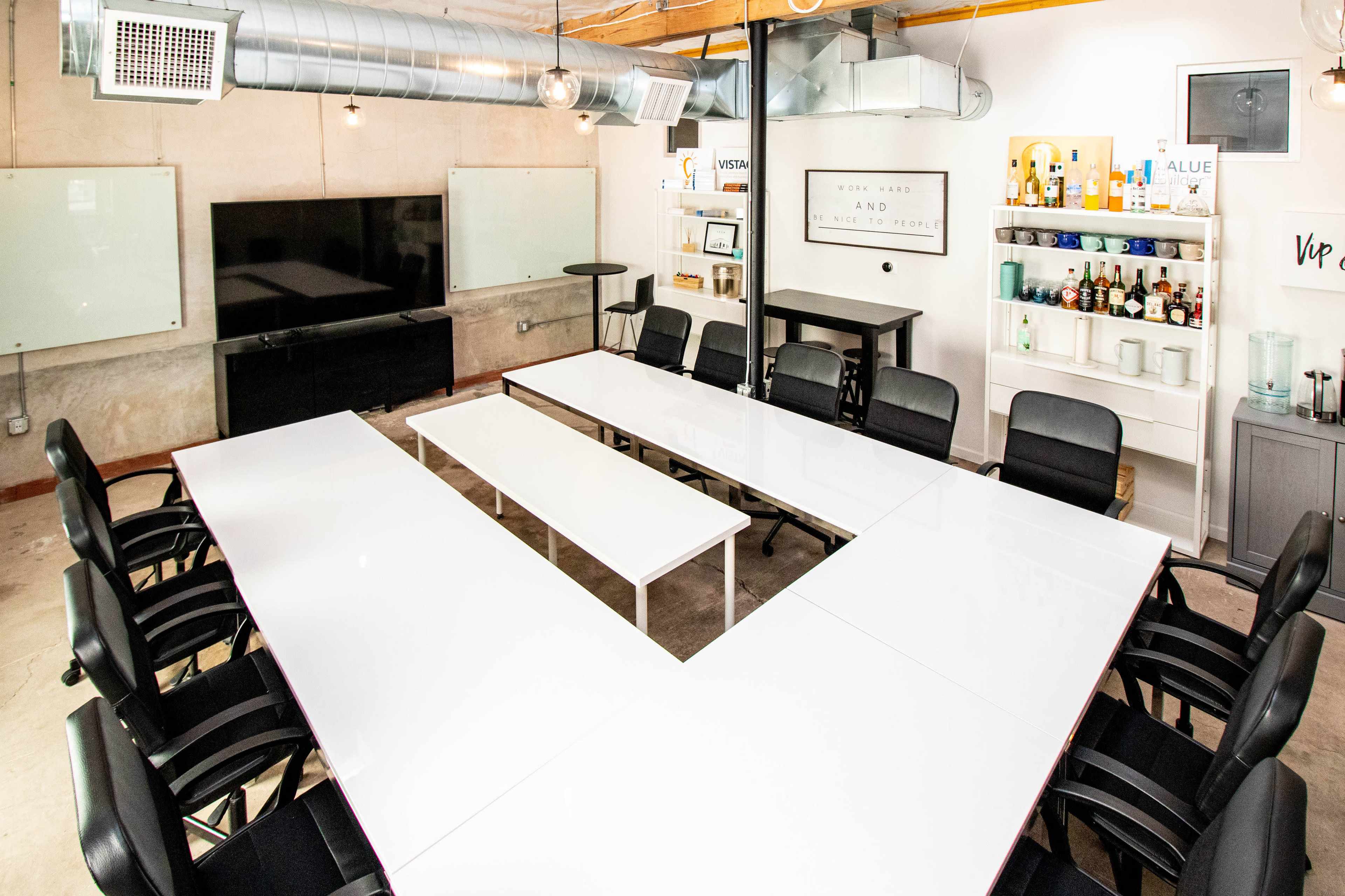 A modern conference room features a large white table arranged in a U-shape, surrounded by black chairs, with a TV display, whiteboards, and a bar cart in the background.