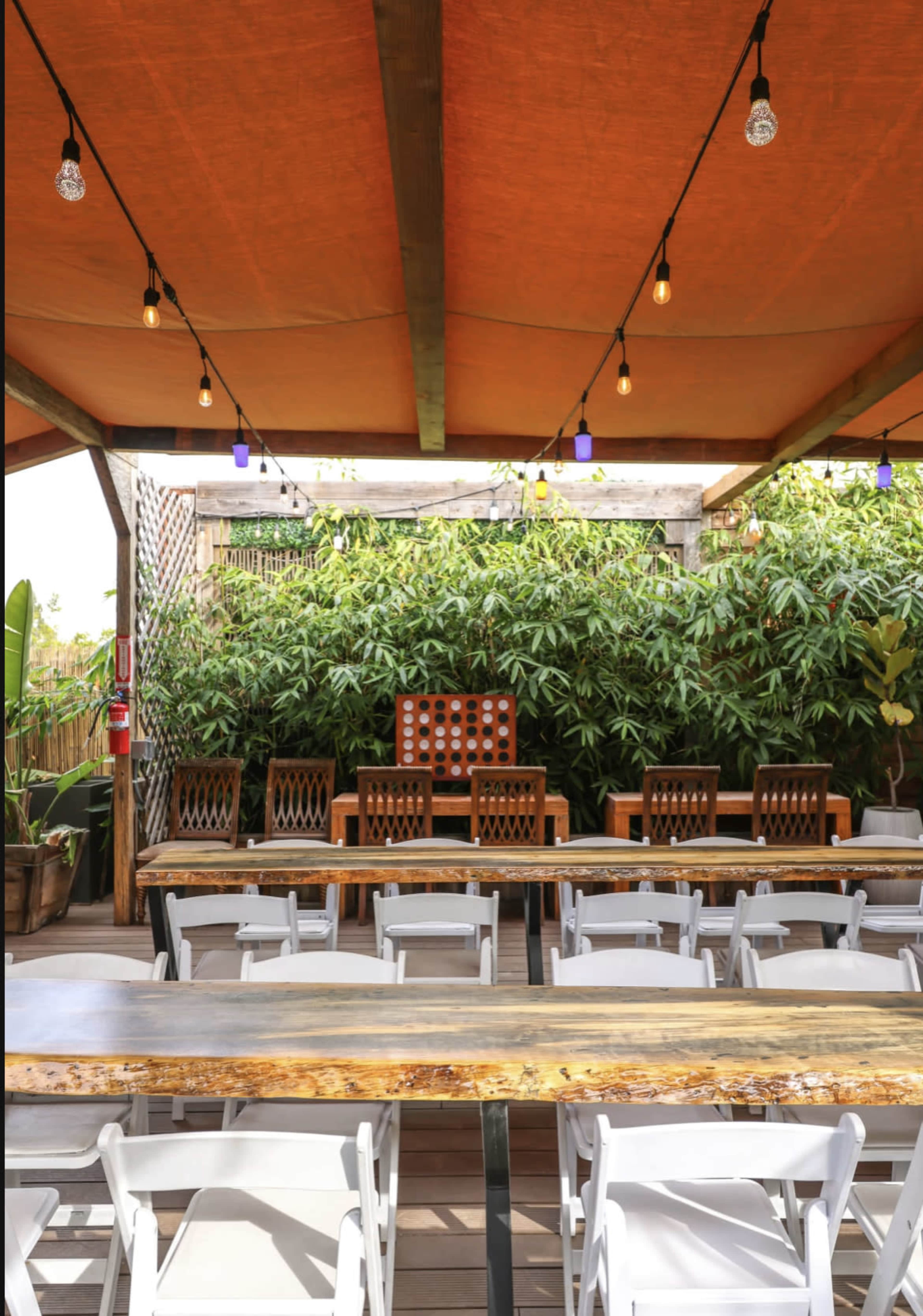 The image shows an outdoor dining area with a long wooden table, white chairs arranged in rows, and a backdrop of lush greenery.