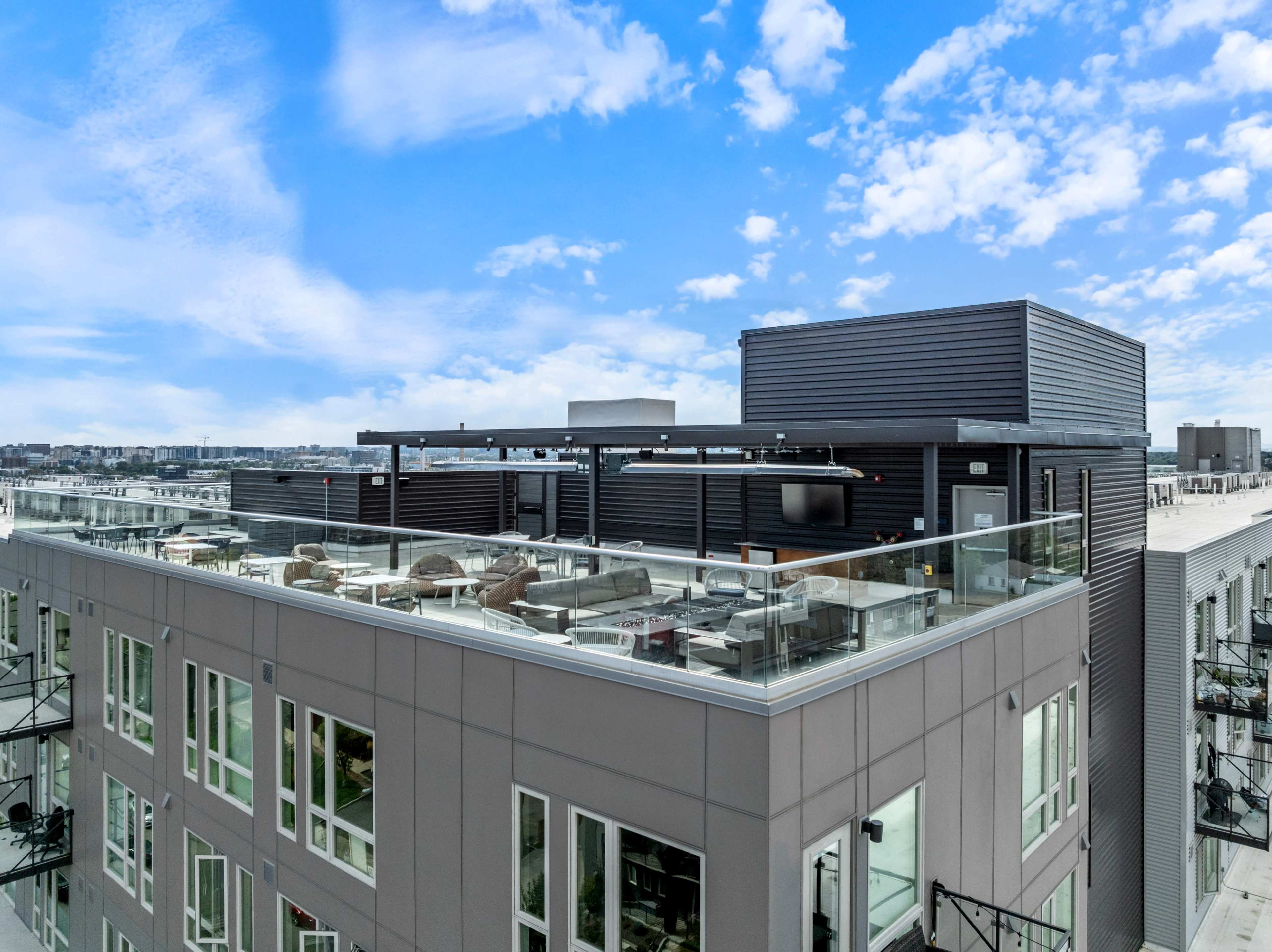 The image shows a modern rooftop terrace atop a multi-story building, featuring seating areas and an outdoor TV, with a clear blue sky in the background.