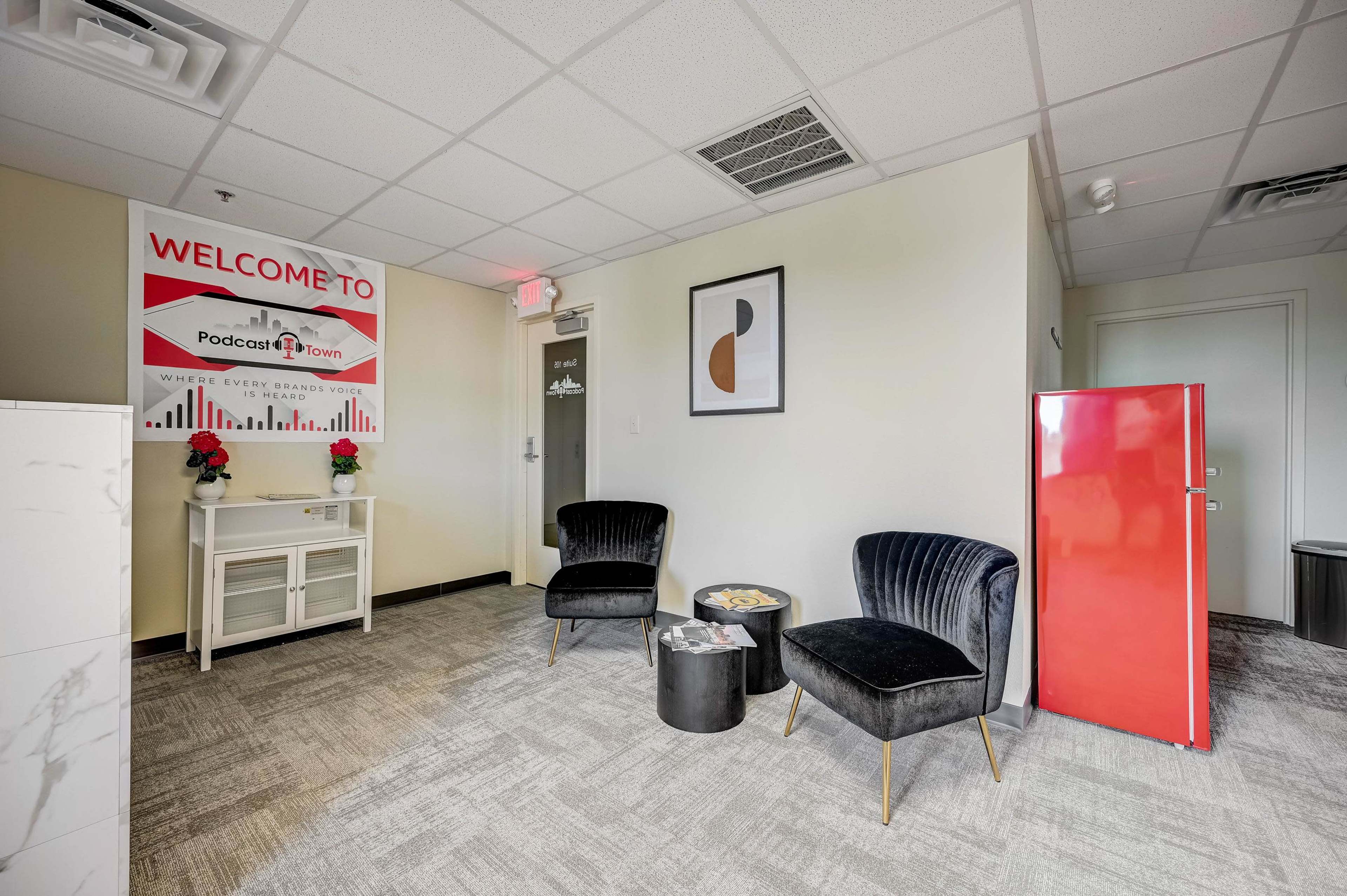 A modern office space featuring two black chairs, a small black table, a red fridge, and a sign that says "Welcome to PodcastTown."