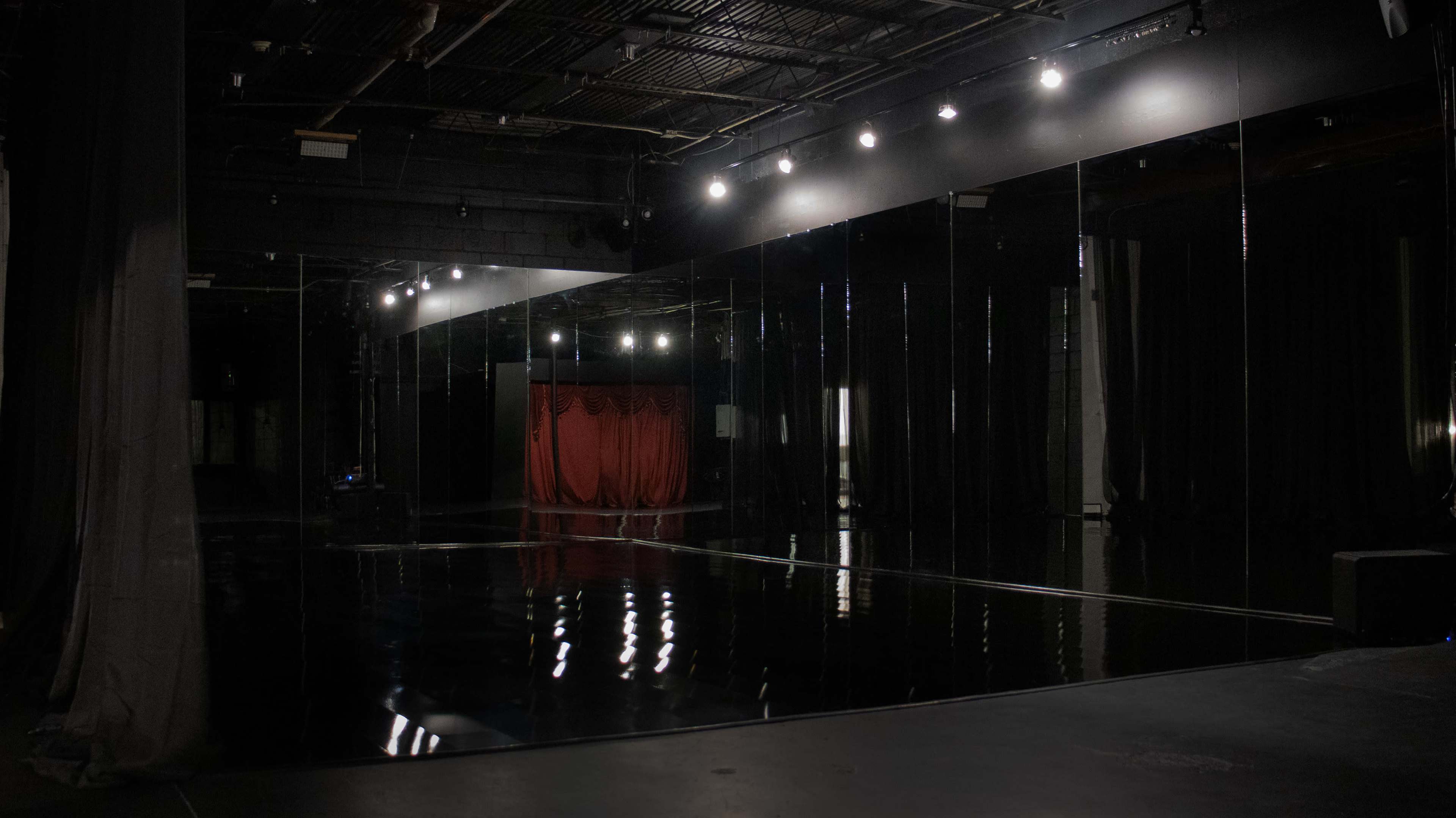 The image shows a dimly lit dance studio with mirrored walls and a red curtain in the background.
