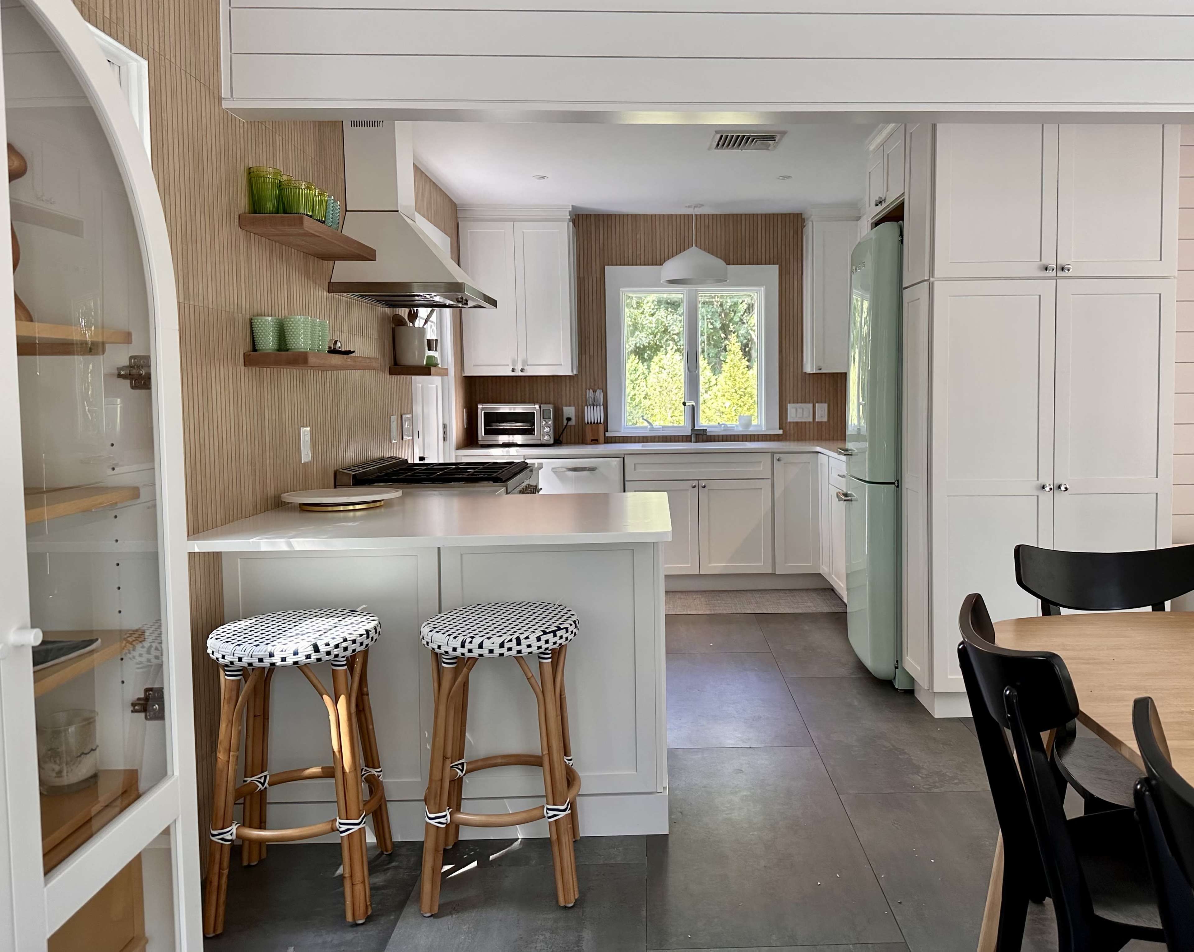 A modern kitchen features white cabinetry, a light wooden accent wall, and a dining area with black chairs and two patterned stools.