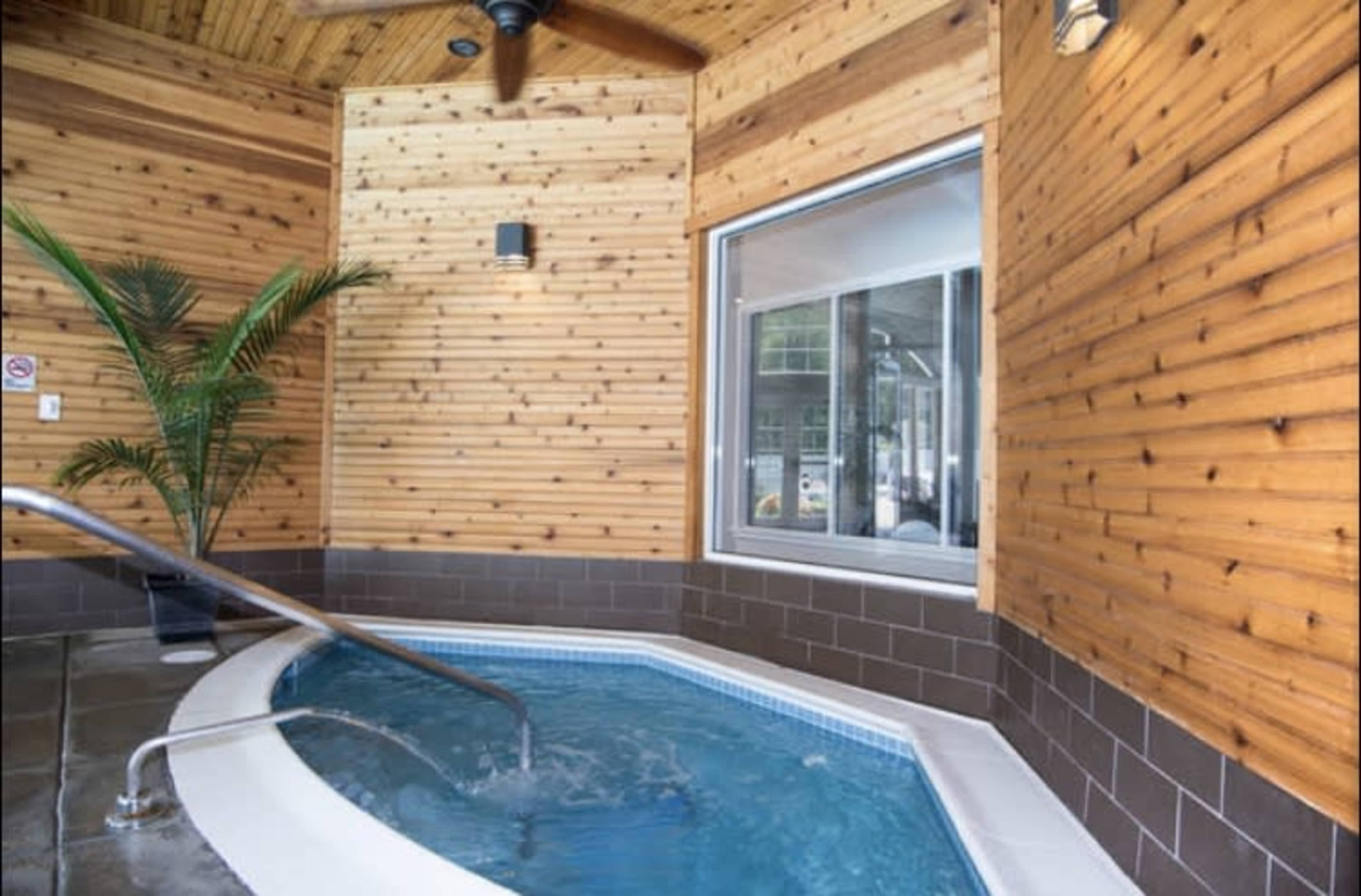 The image shows a small indoor spa area with a circular hot tub, wooden walls, and a large window.