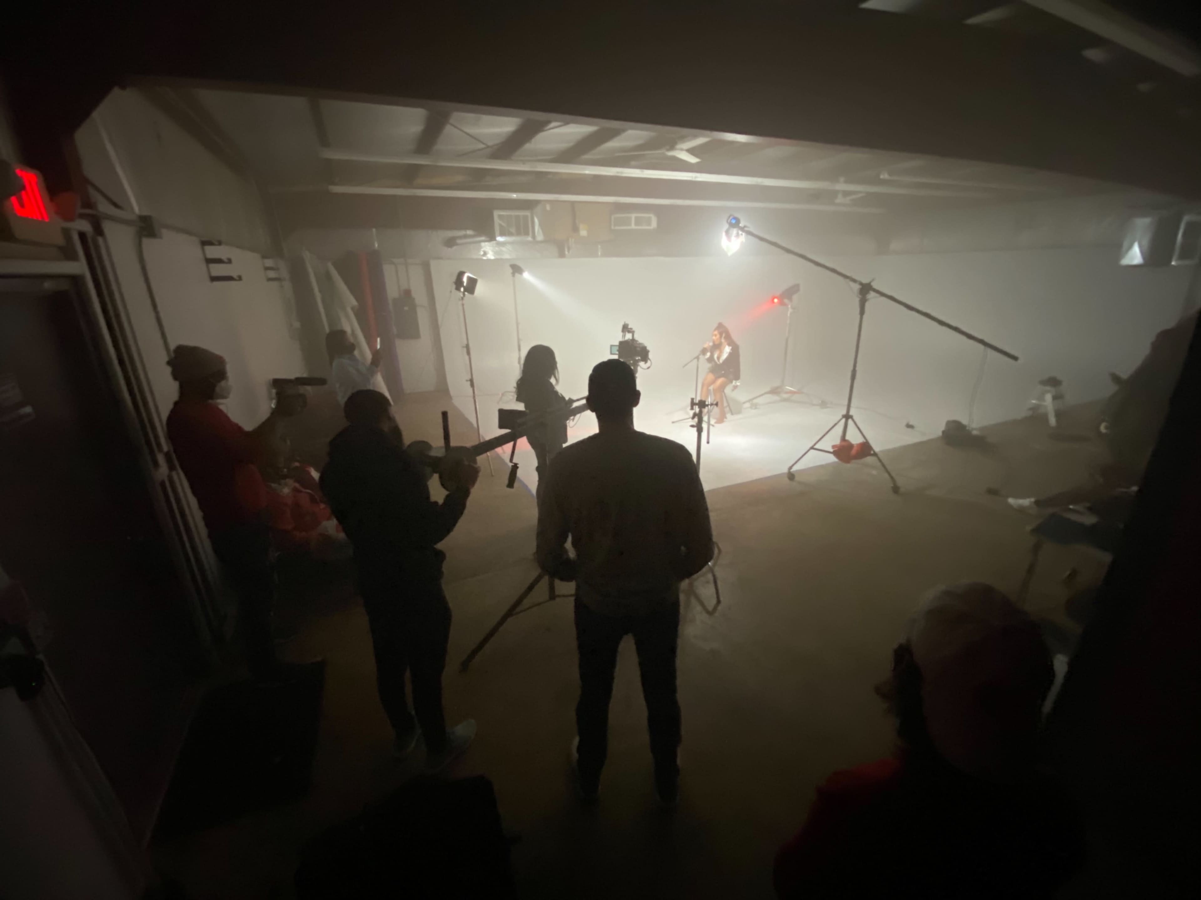 A group of individuals stands in a dimly lit room, observing a subject illuminated by lights on a set.