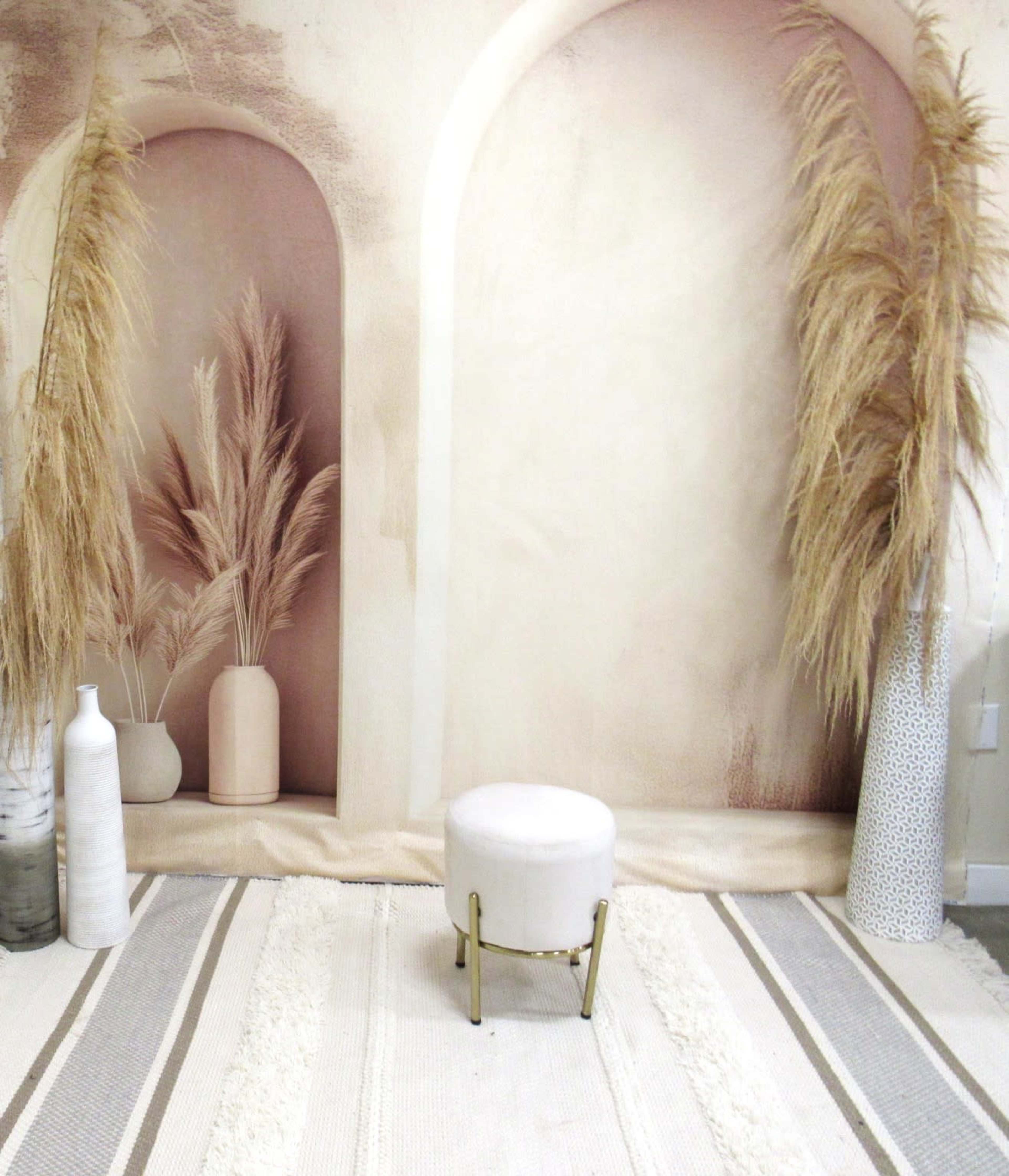 The image features a simple interior scene with a white stool in front of a backdrop depicting two archways, flanked by ornamental grasses in vases.