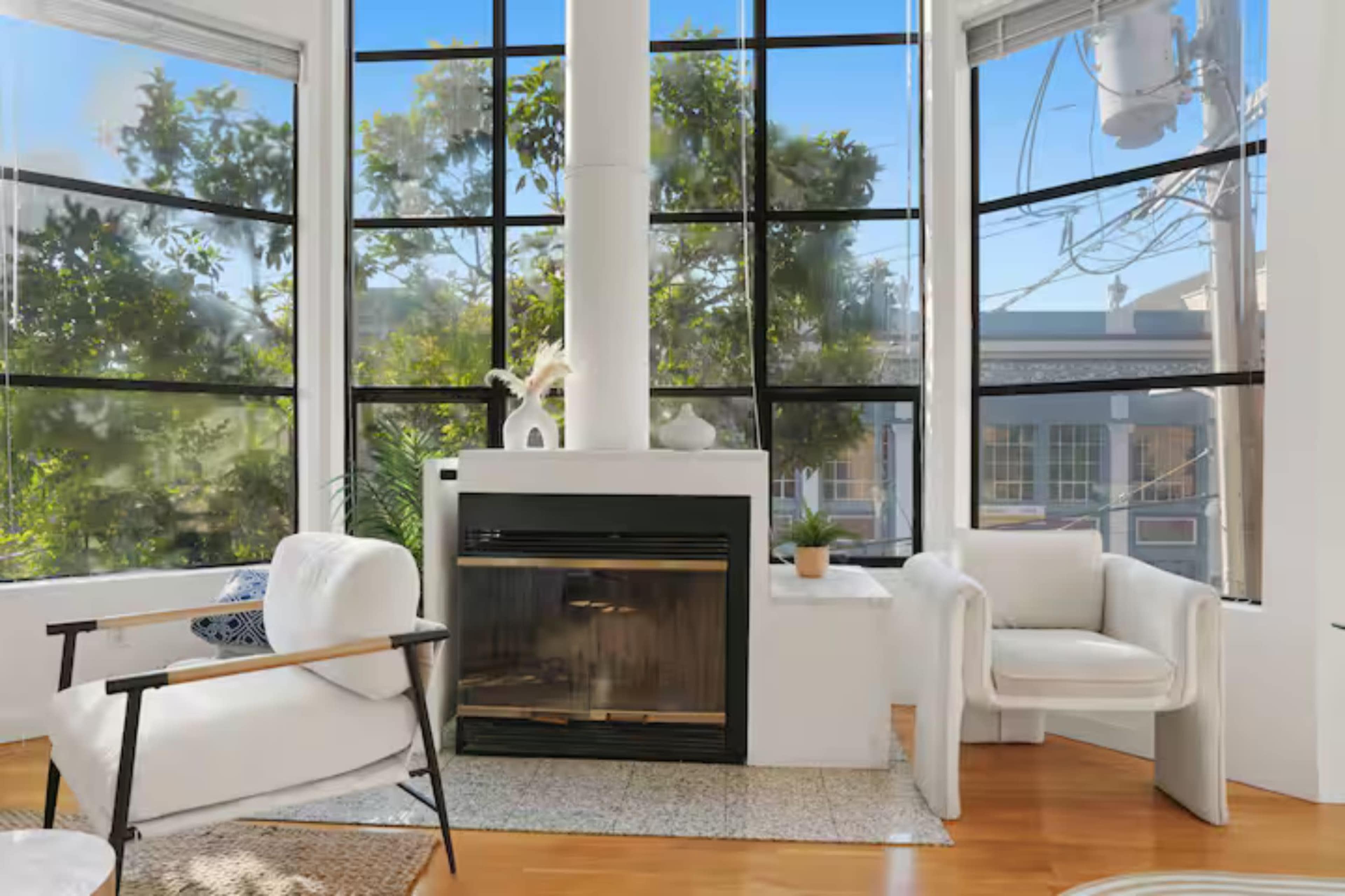 The image shows a bright living space with large windows, a white fireplace, and modern seating arrangements.