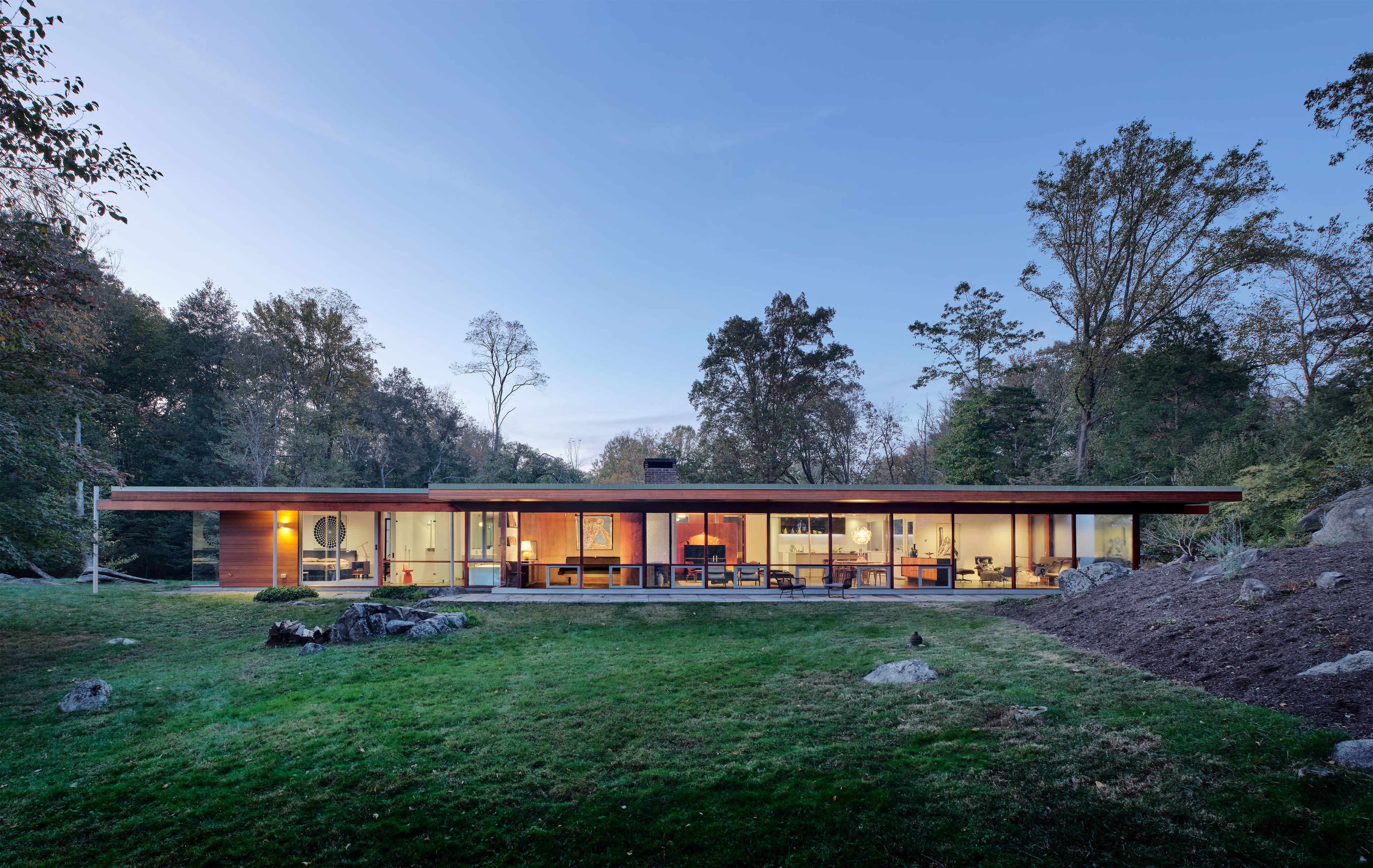 A single-story modern house with large glass windows is surrounded by trees and a grassy lawn.