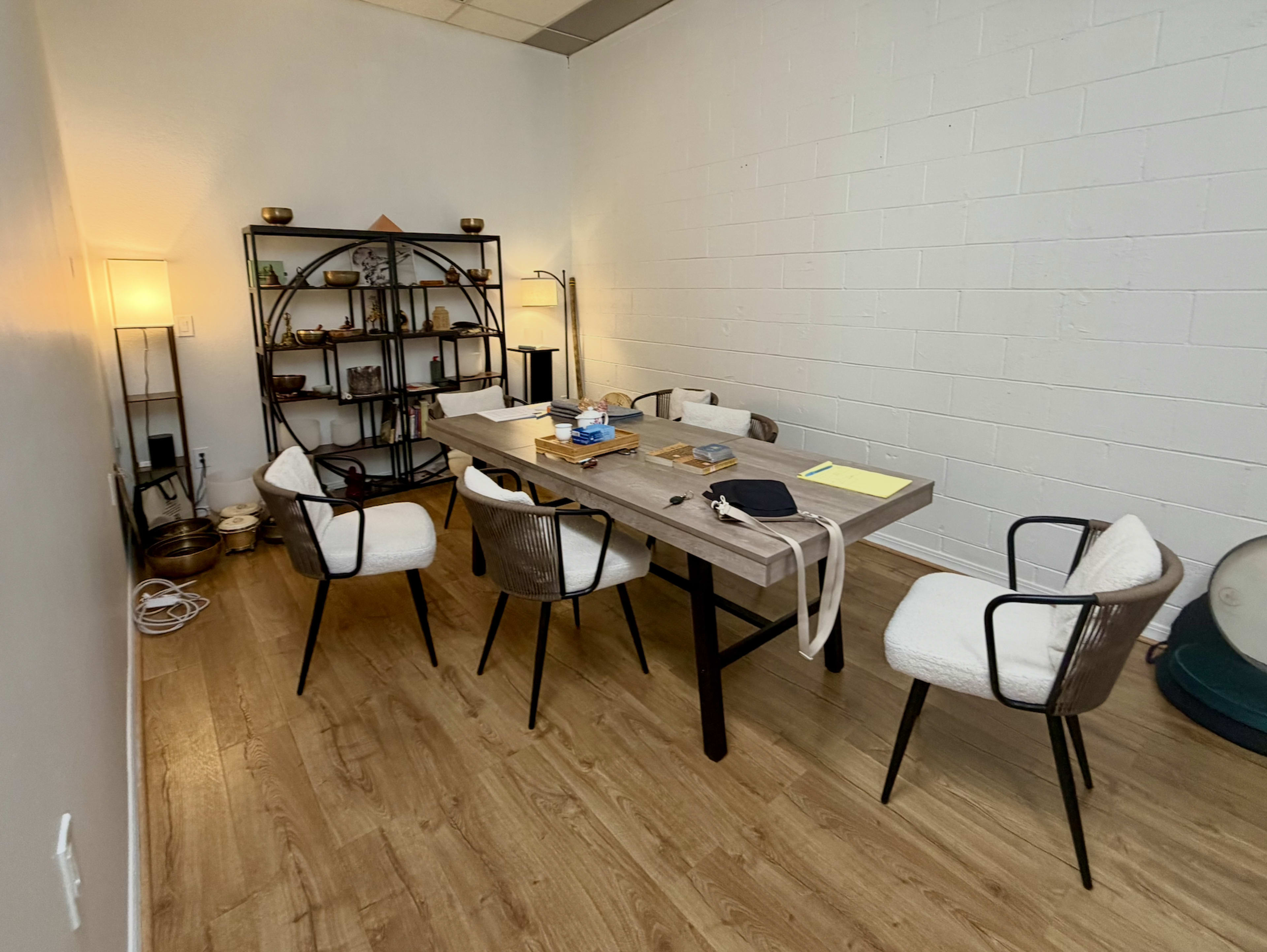 The image shows a small meeting room with a wooden table surrounded by four chairs, a bookcase with items on display, and a lamp in the corner.