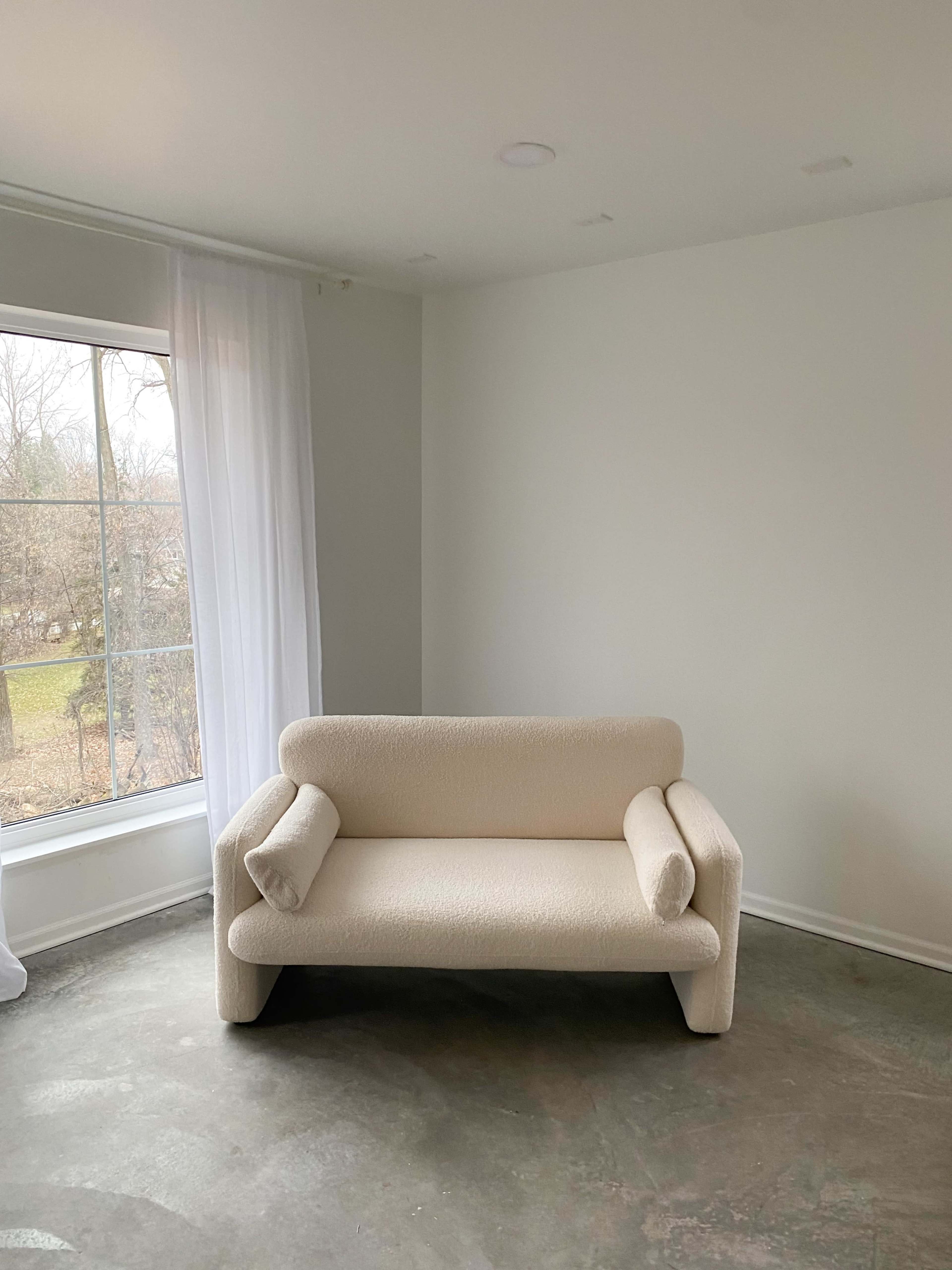 A beige, plush sofa with rounded arms sits in a minimalist room featuring a large window with sheer white curtains.