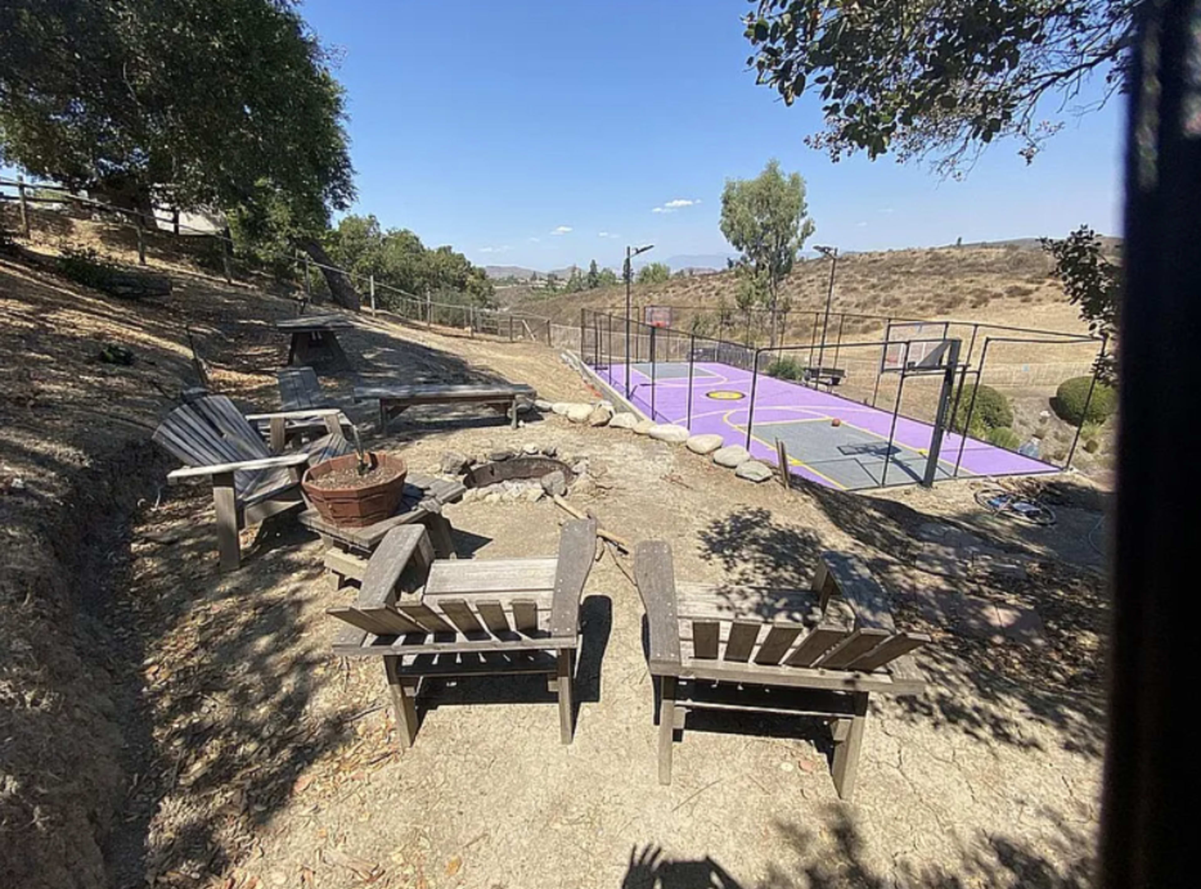 The image shows a hillside area with two wooden chairs facing a fire pit, overlooking a purple multi-use sports court surrounded by trees and a dry landscape.