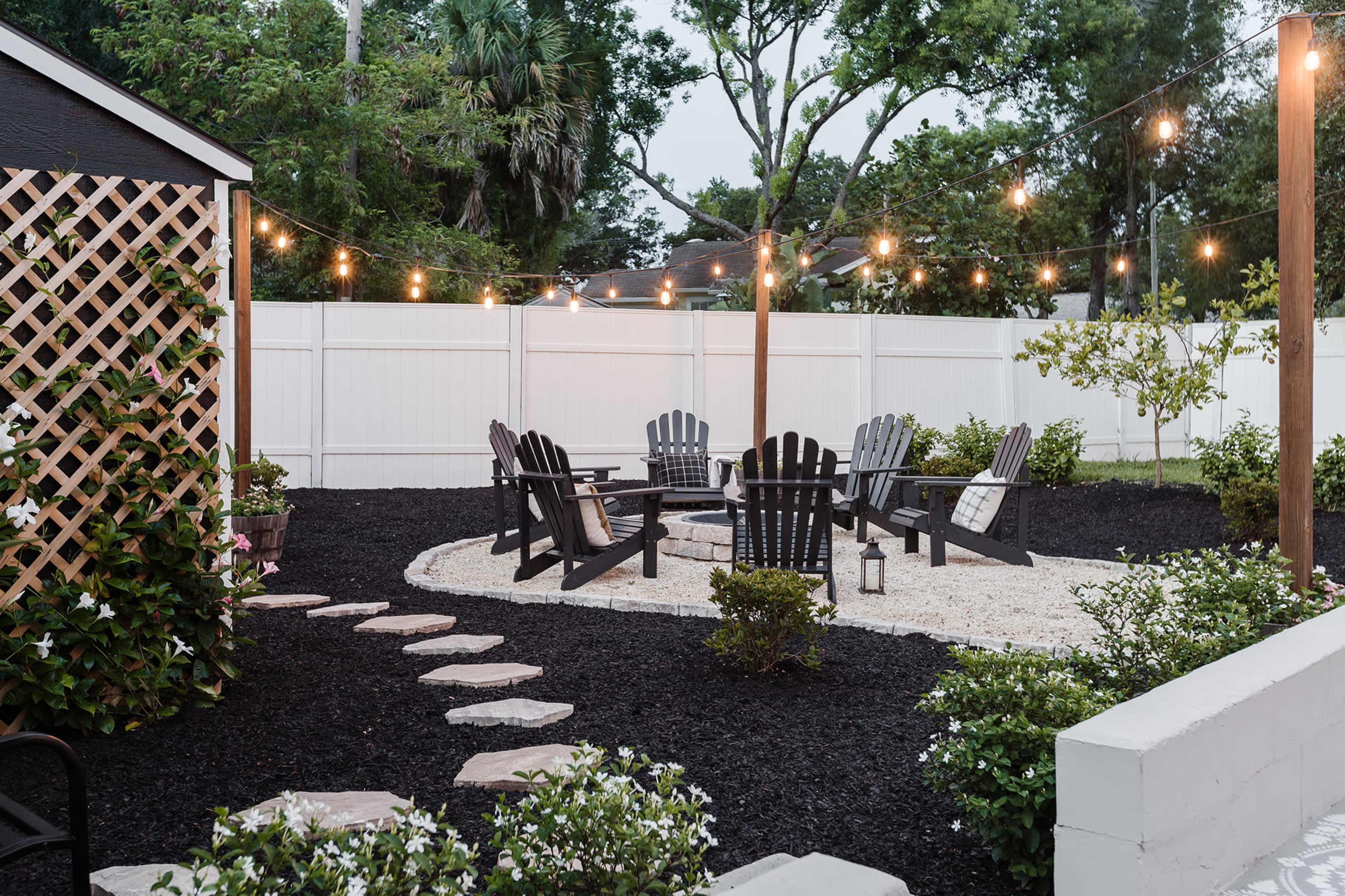 A circular seating area with Adirondack chairs is surrounded by decorative gravel and lined with stepping stones, illuminated by string lights in a landscaped backyard setting.