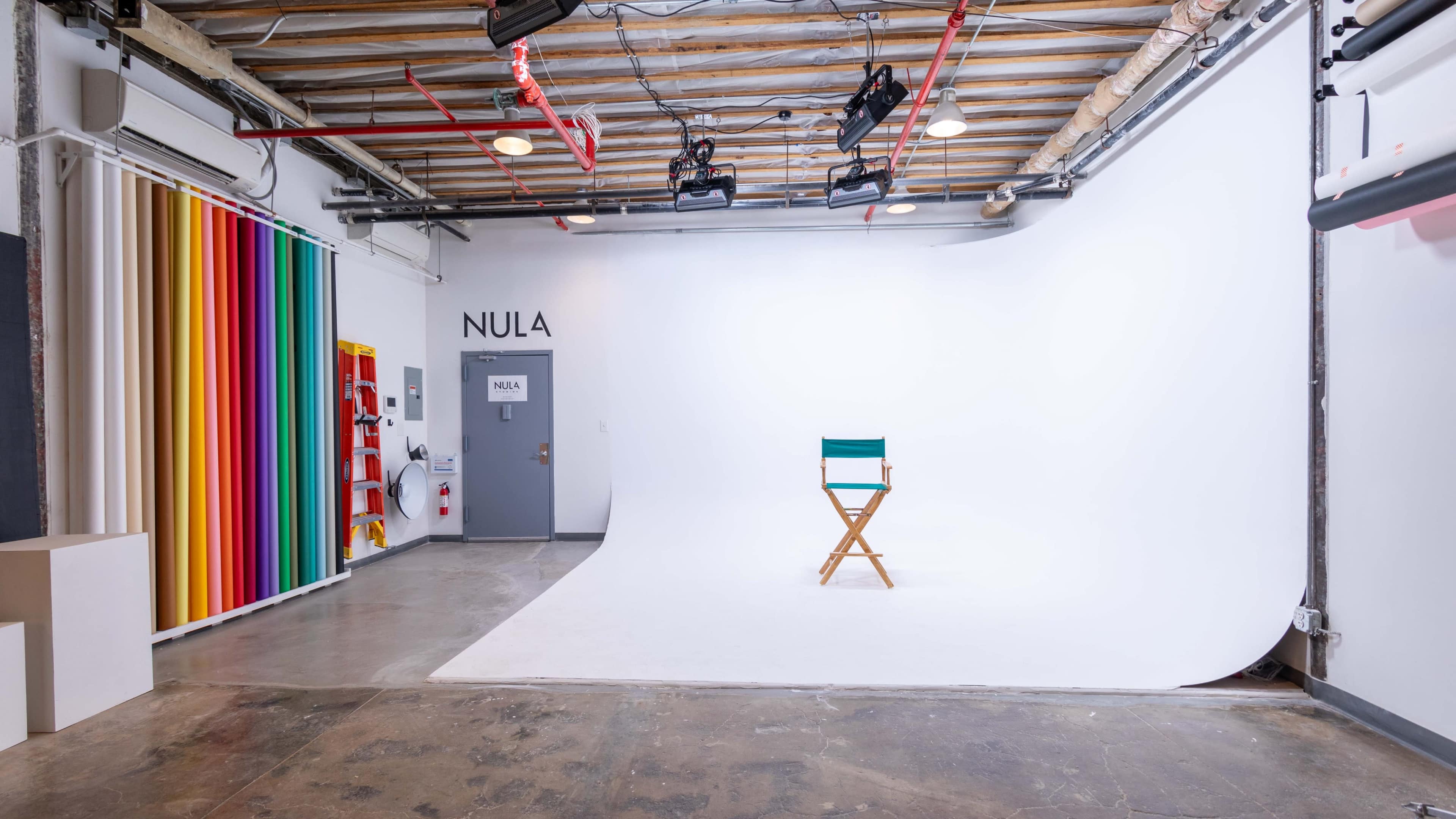 A minimalist photo studio with a white backdrop, a wooden director's chair in the center, and colorful striped panels on the side walls.