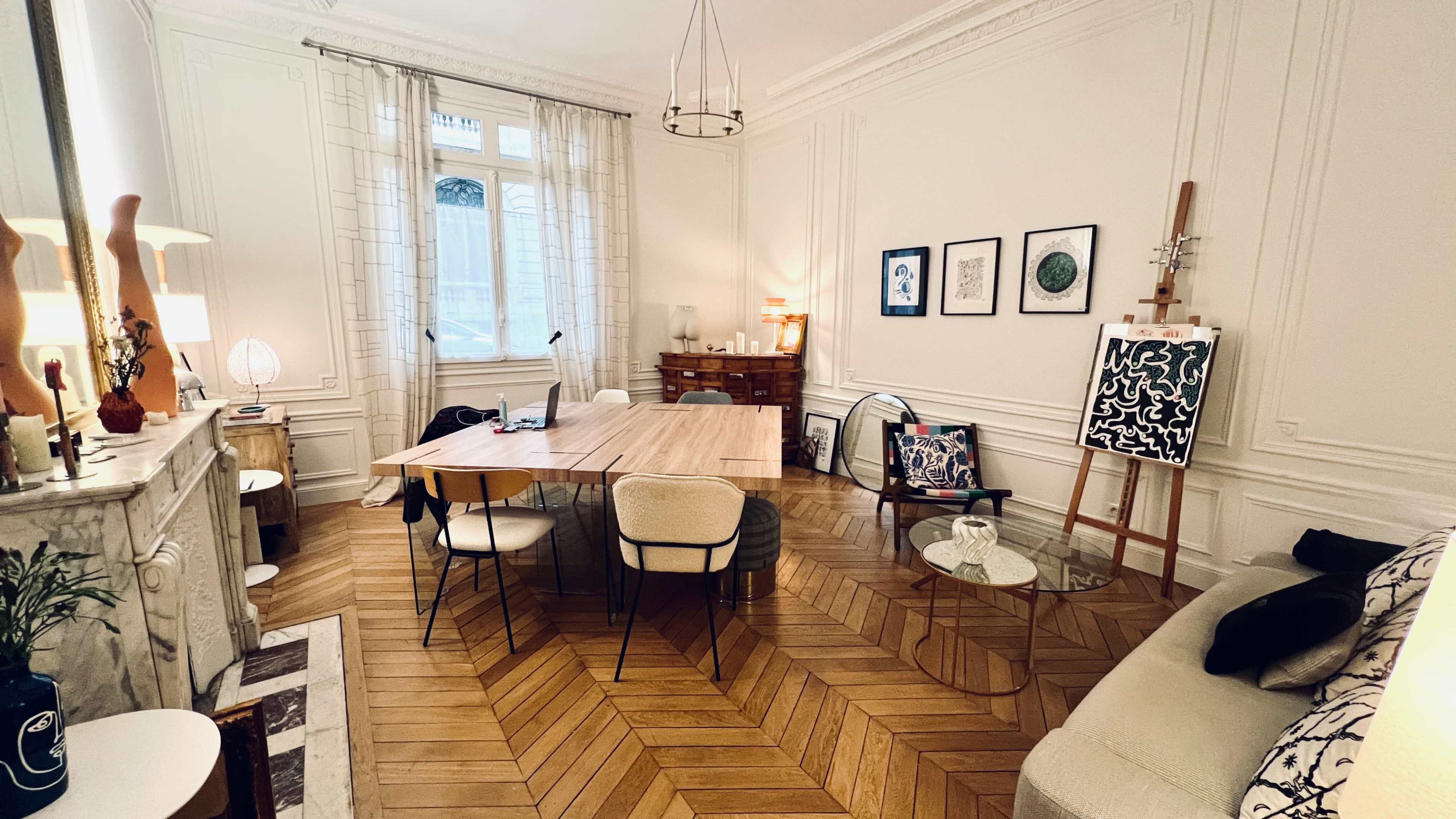 The image shows a bright, elegantly decorated living space with a wooden dining table, various chairs, a sofa, and framed artwork on the walls.