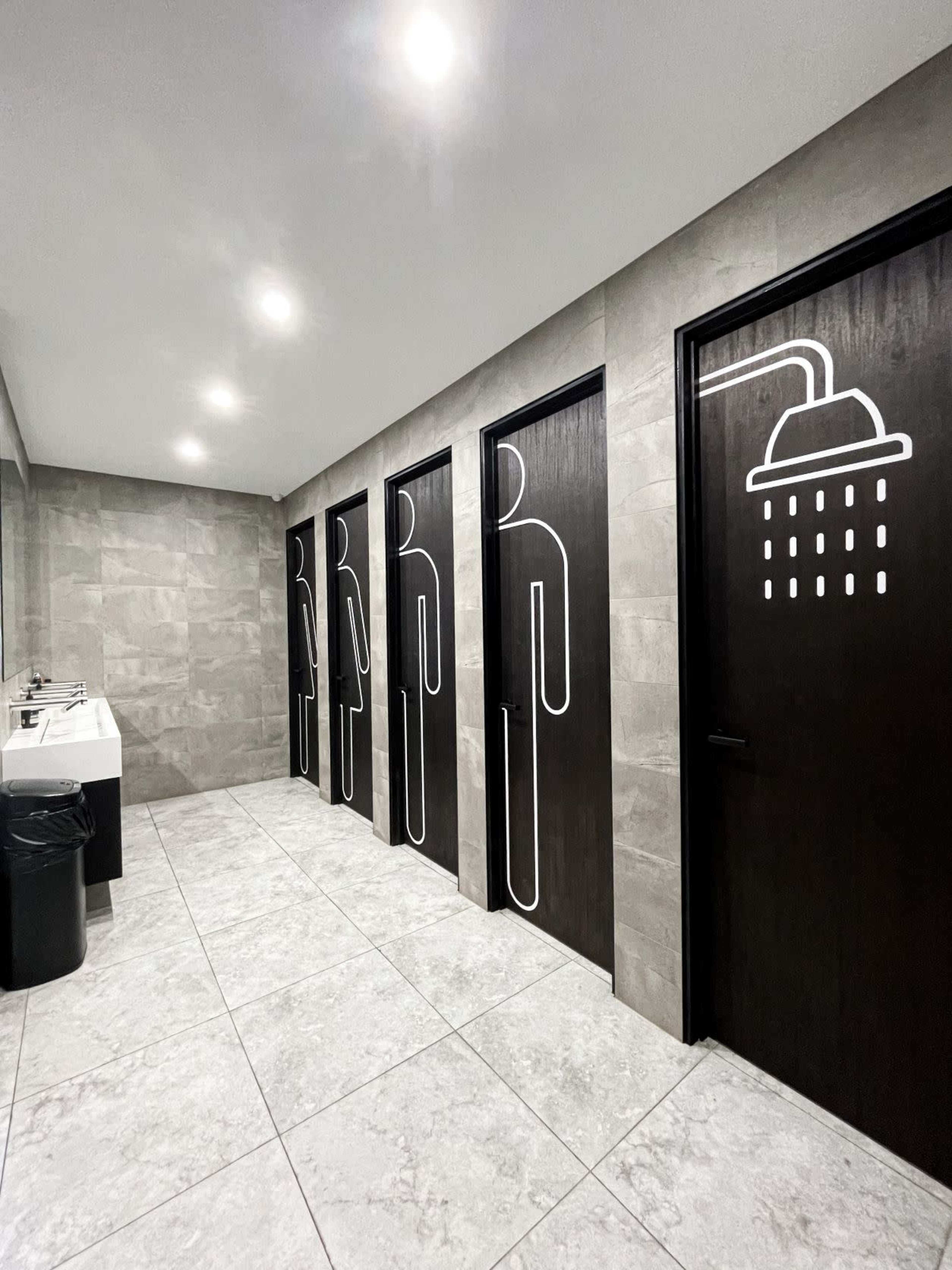 A modern restroom features four doors with minimalist symbols indicating men's and women's facilities and a shower area, alongside a sink and trash bin.