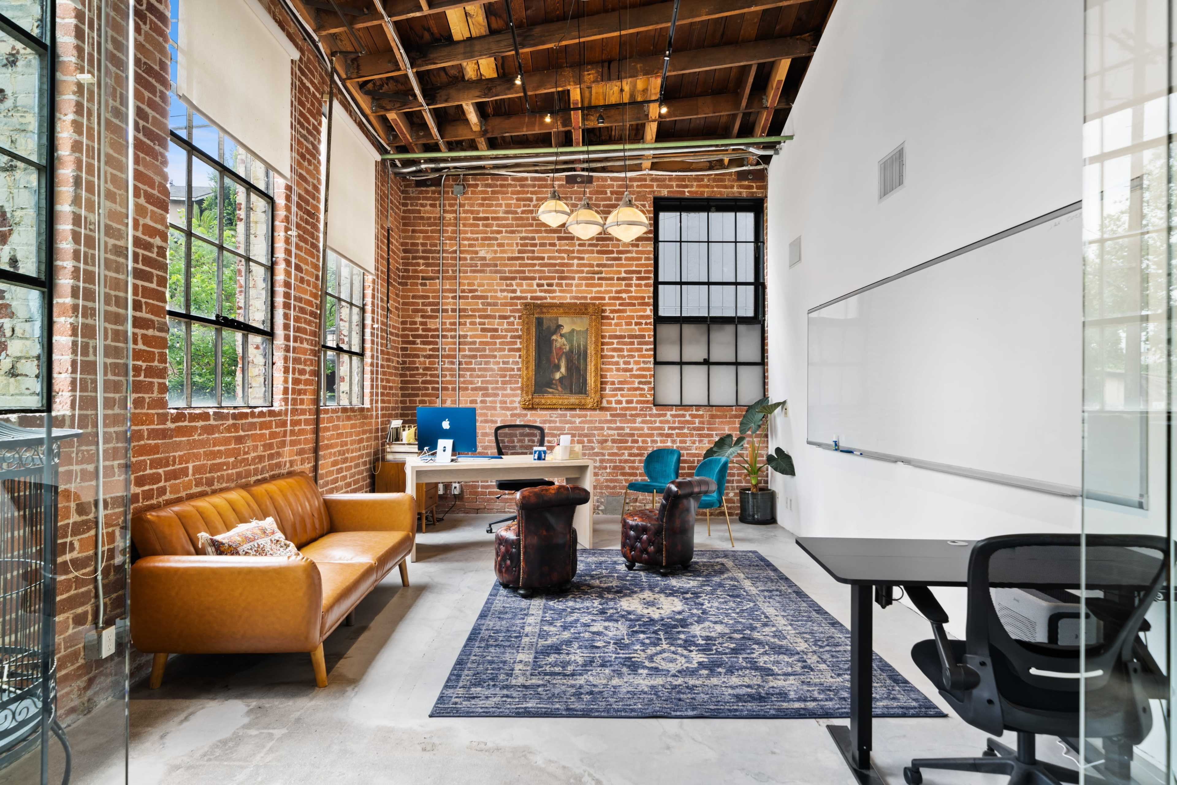 A modern office space featuring exposed brick walls, wooden beams, a large window, and various furnishings including a brown leather sofa and blue chairs.