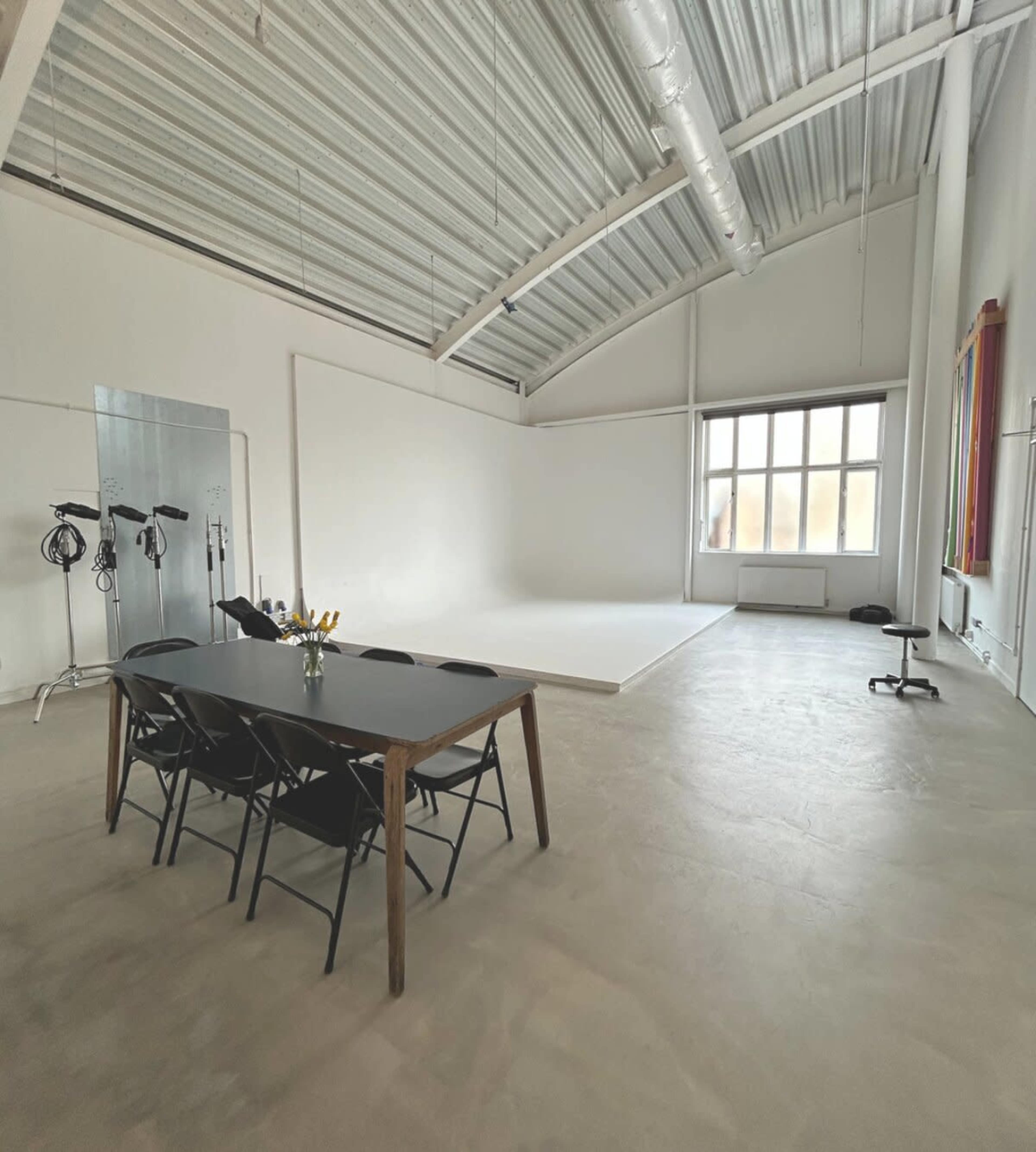 The image shows a spacious, well-lit studio with a large white wall, a small white platform, and a black table with chairs.