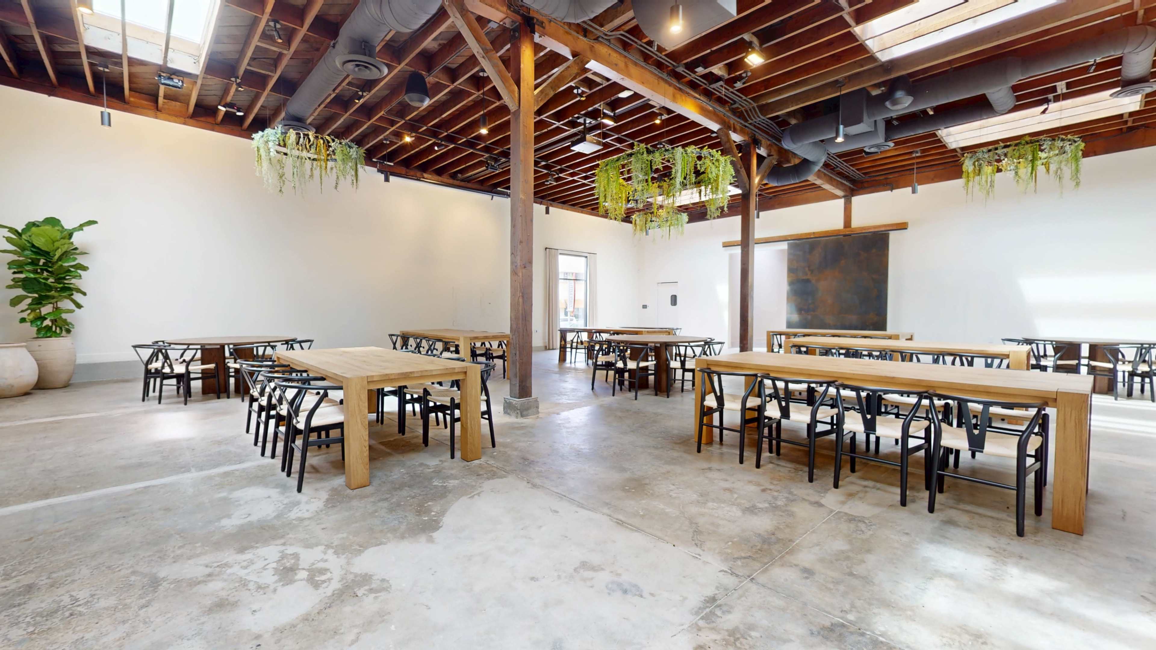 The image depicts a spacious, well-lit room with wooden beams, multiple long tables arranged for seating, and hanging greenery on the ceiling.