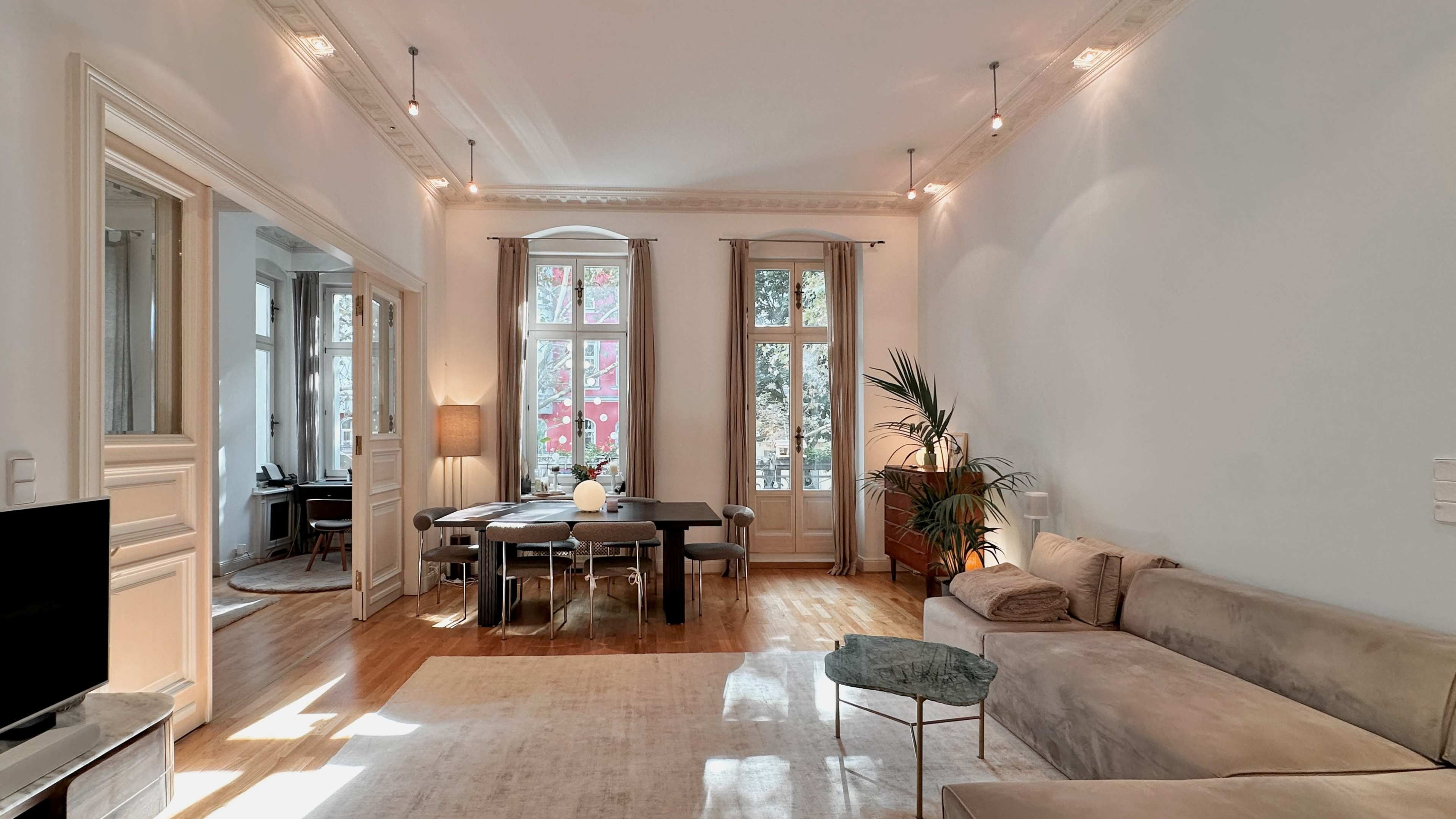 The image shows a bright, spacious living room with large windows, a dining table, a sectional couch, and a potted plant.