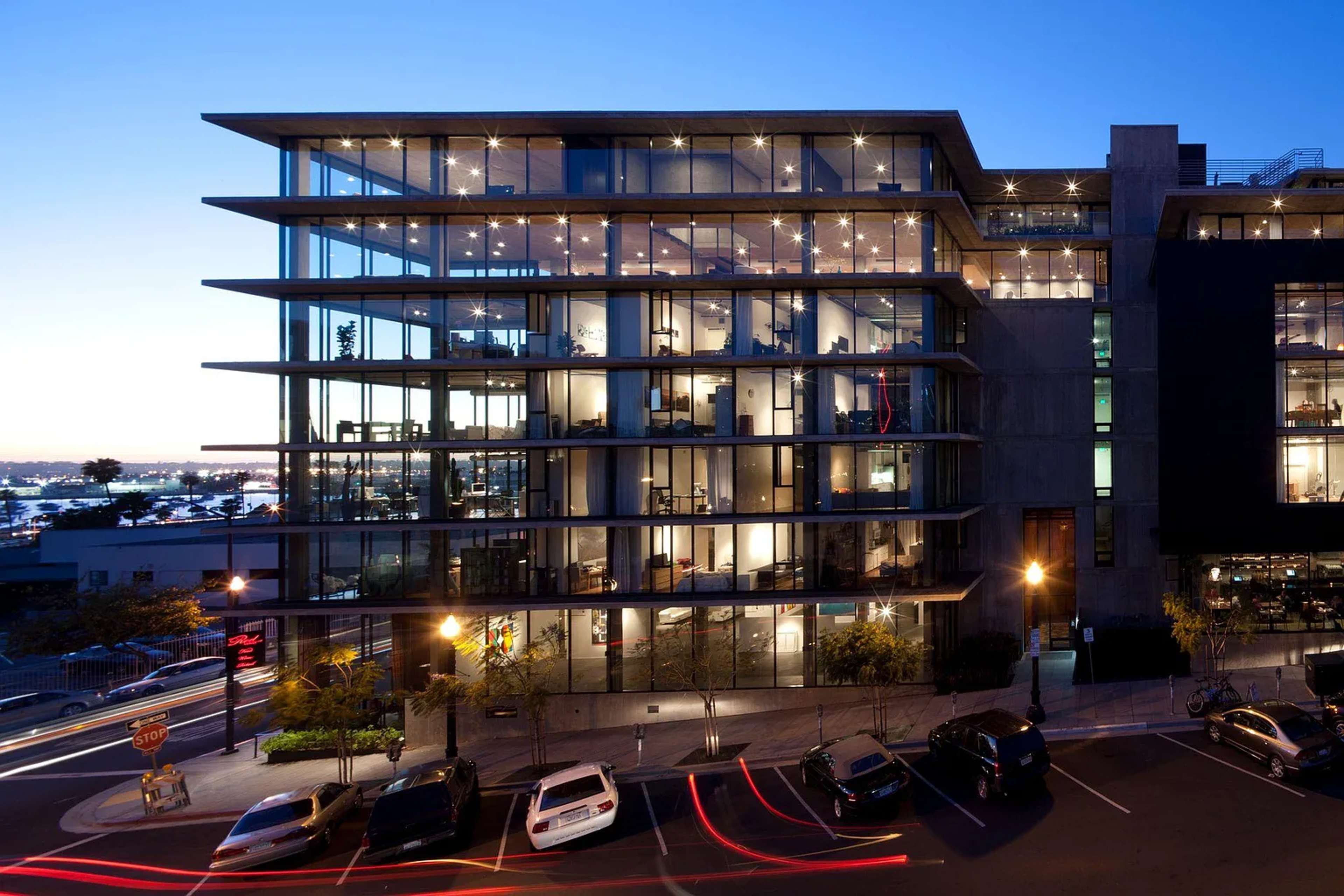 The image shows a modern multi-story building with a glass façade, illuminated windows, and parked cars on the street below during twilight.