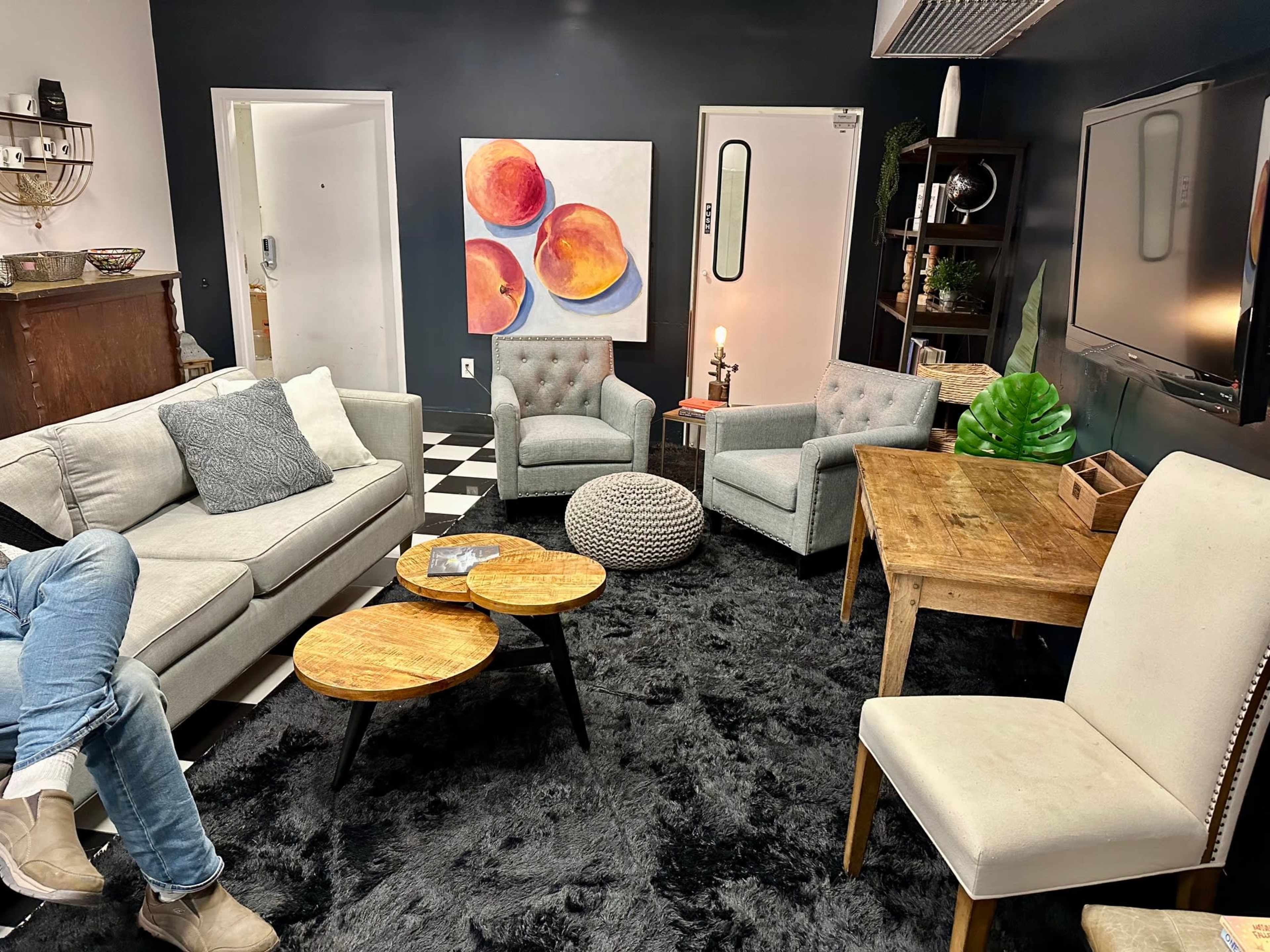 A modern living space featuring a grey sofa, two light grey armchairs, a wooden table, a round pouf, and a black and white checkered floor, with a large artwork of peaches on the wall.