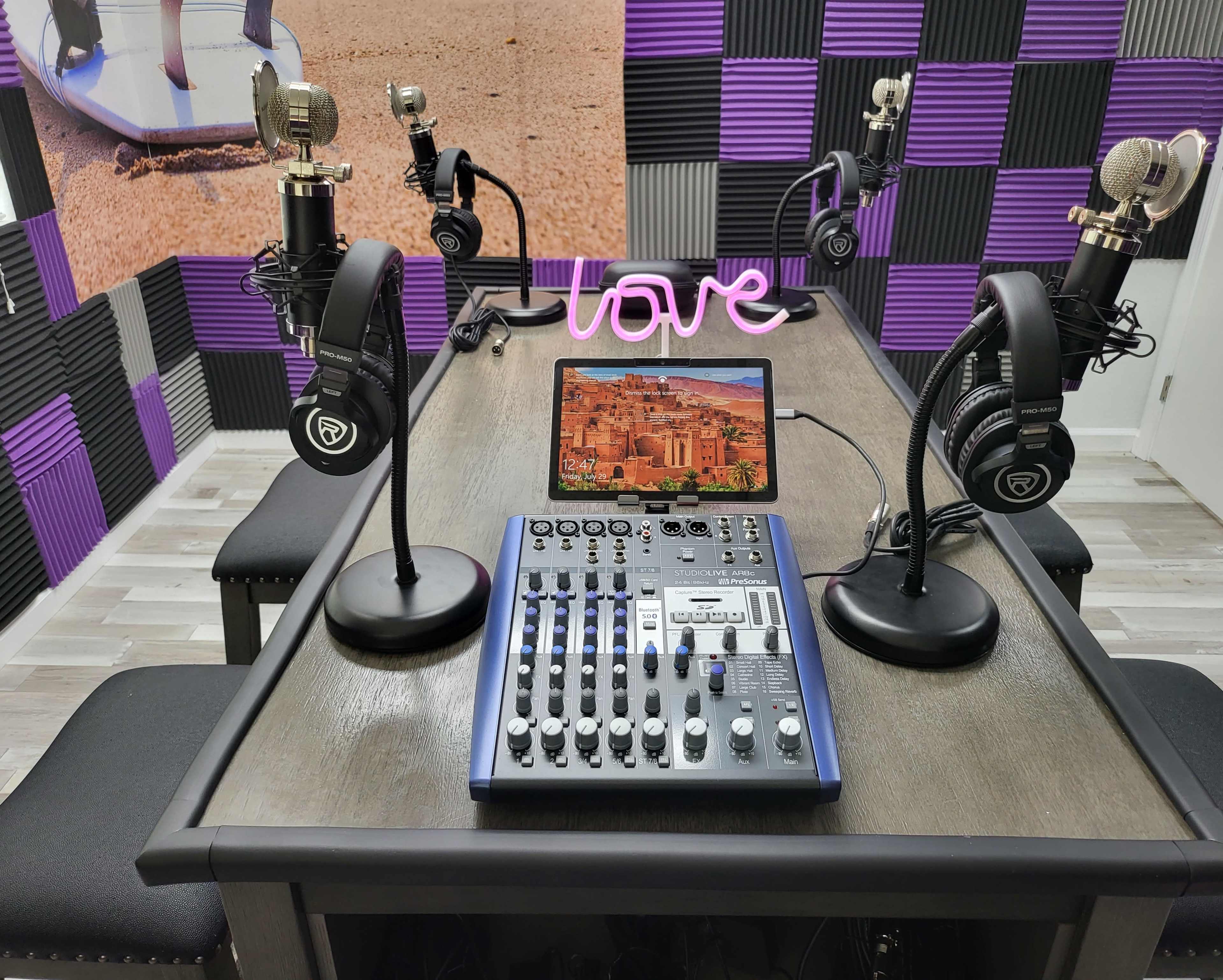 The image shows a recording studio setup with four microphones, headphones, a sound mixer, and a tablet displaying a scenic background on a table.