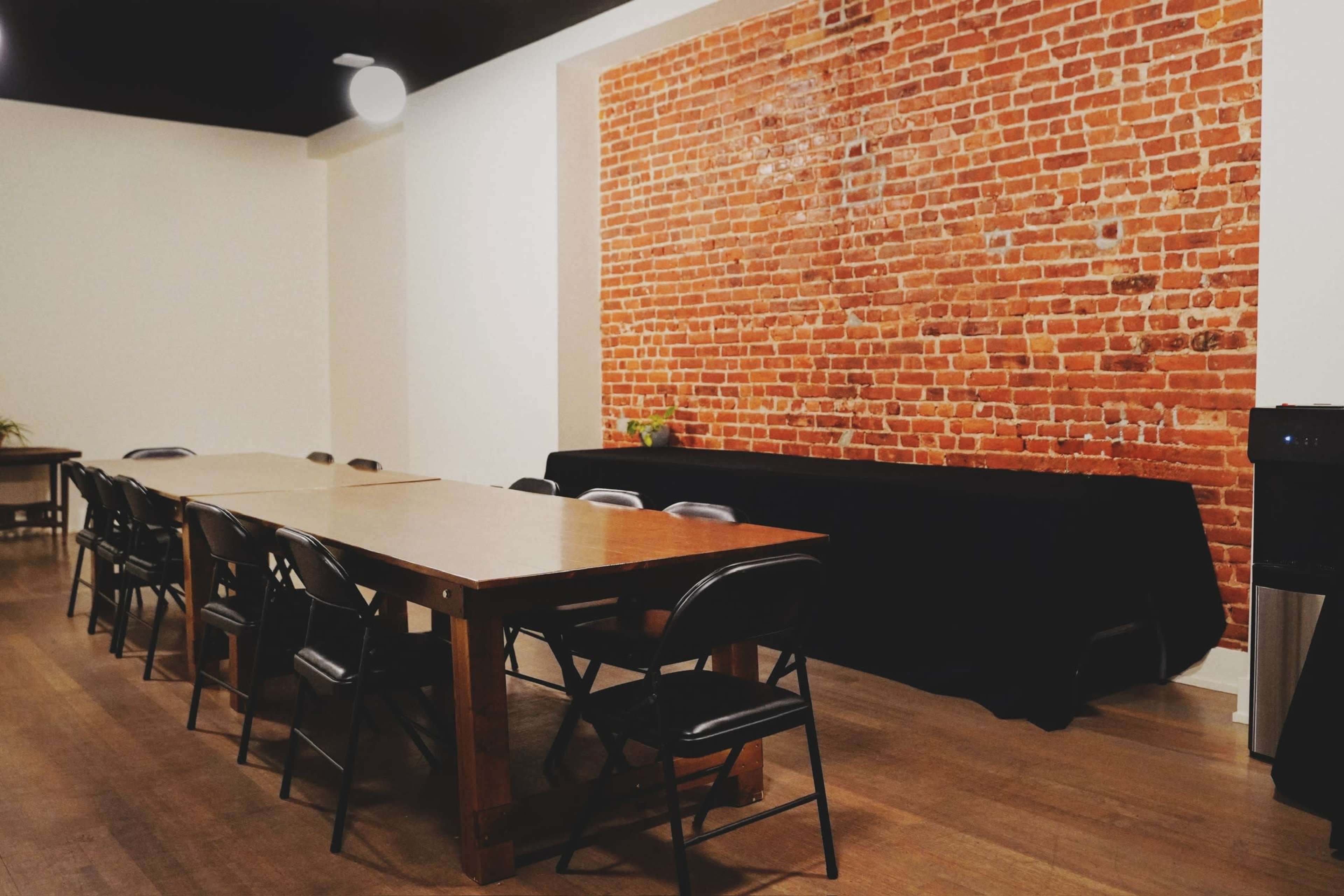 The image shows a spacious room with a long wooden table and black chairs, featuring a exposed brick wall and minimalistic decor.