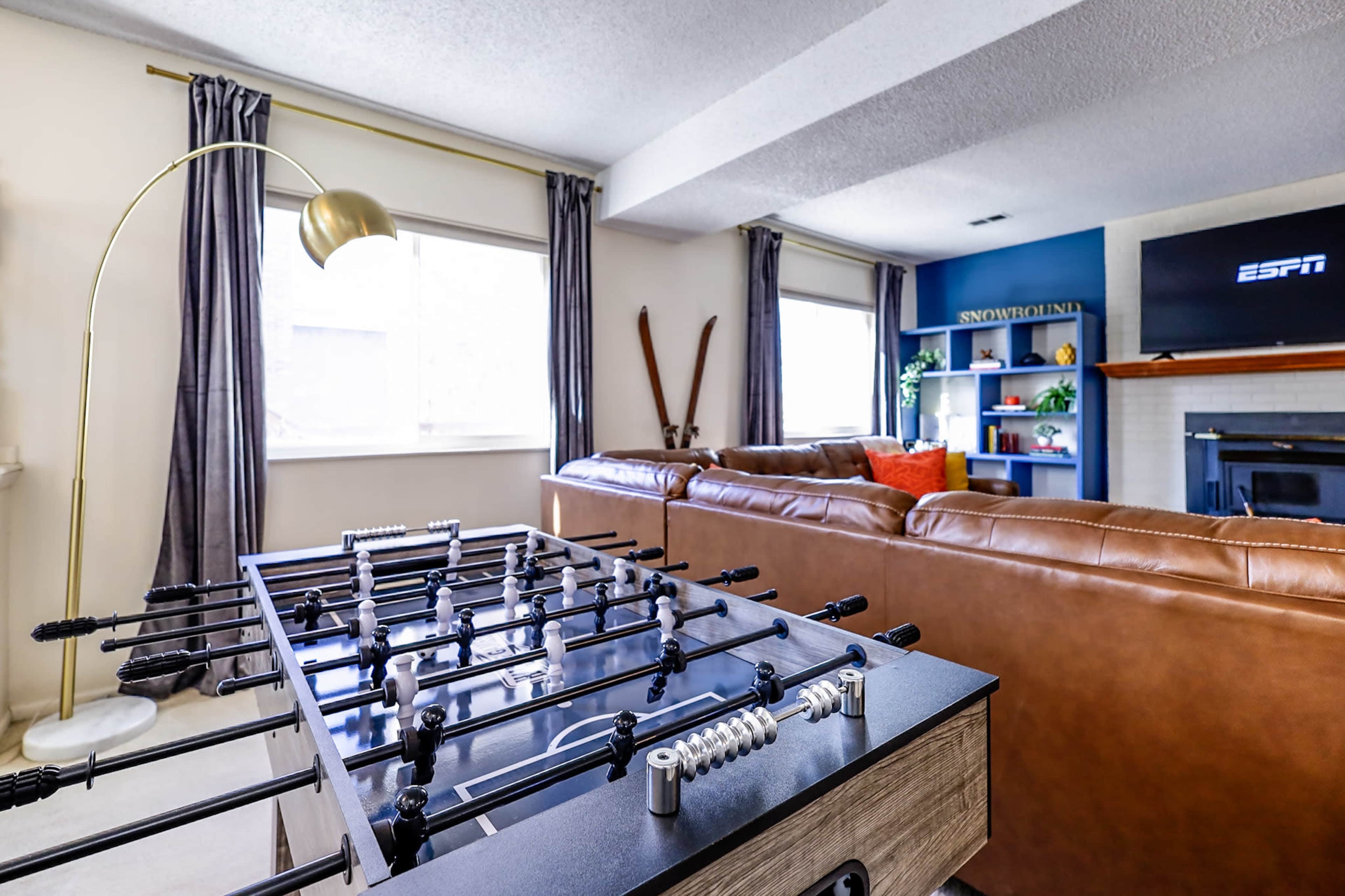 A living room with a foosball table in the foreground, a comfortable seating area with brown leather sofas, and a television mounted on the wall.