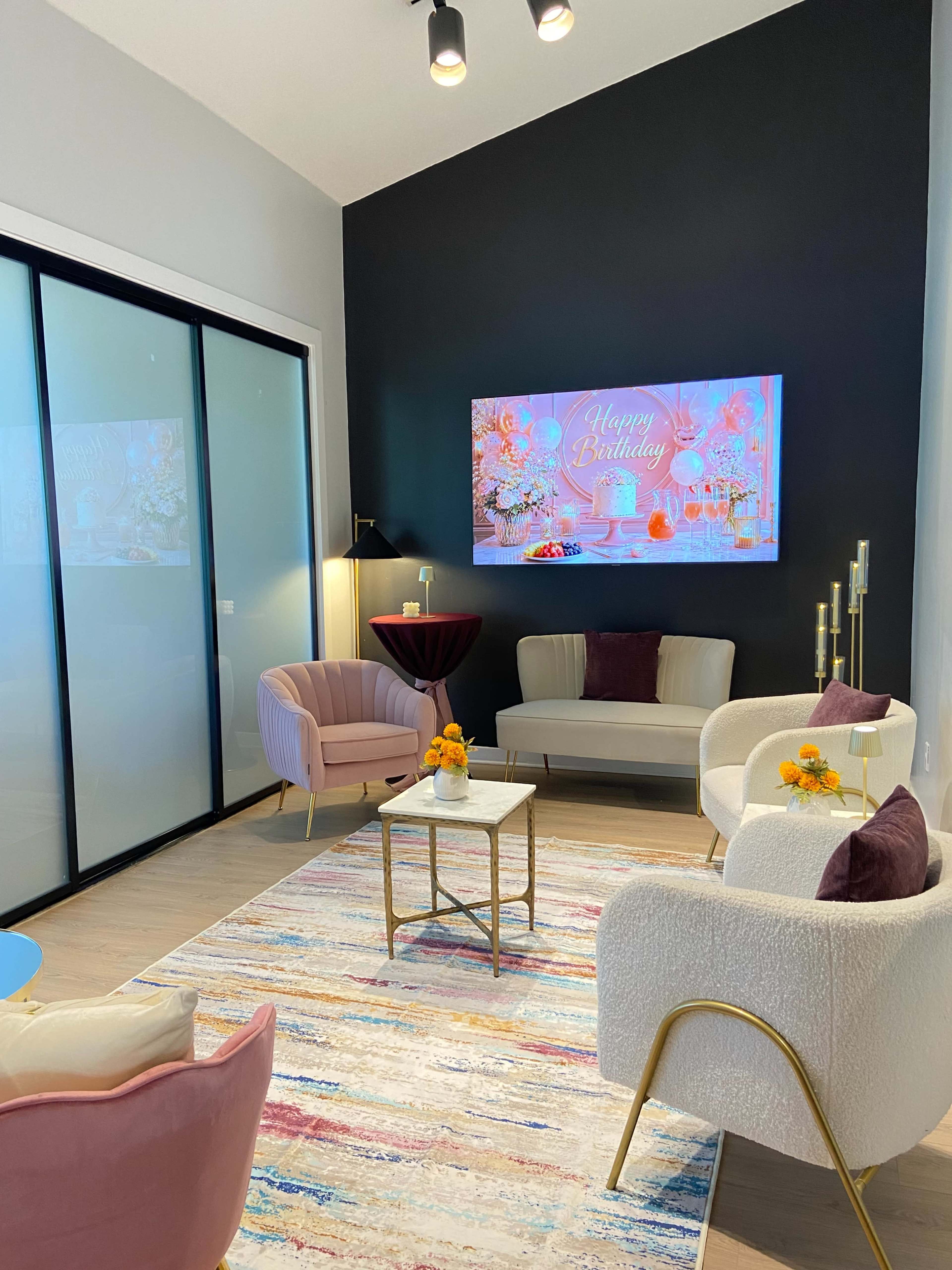 The image depicts a modern living space with a colorful rug, pastel-colored furniture, a wall-mounted birthday greeting, and a small table adorned with flowers.