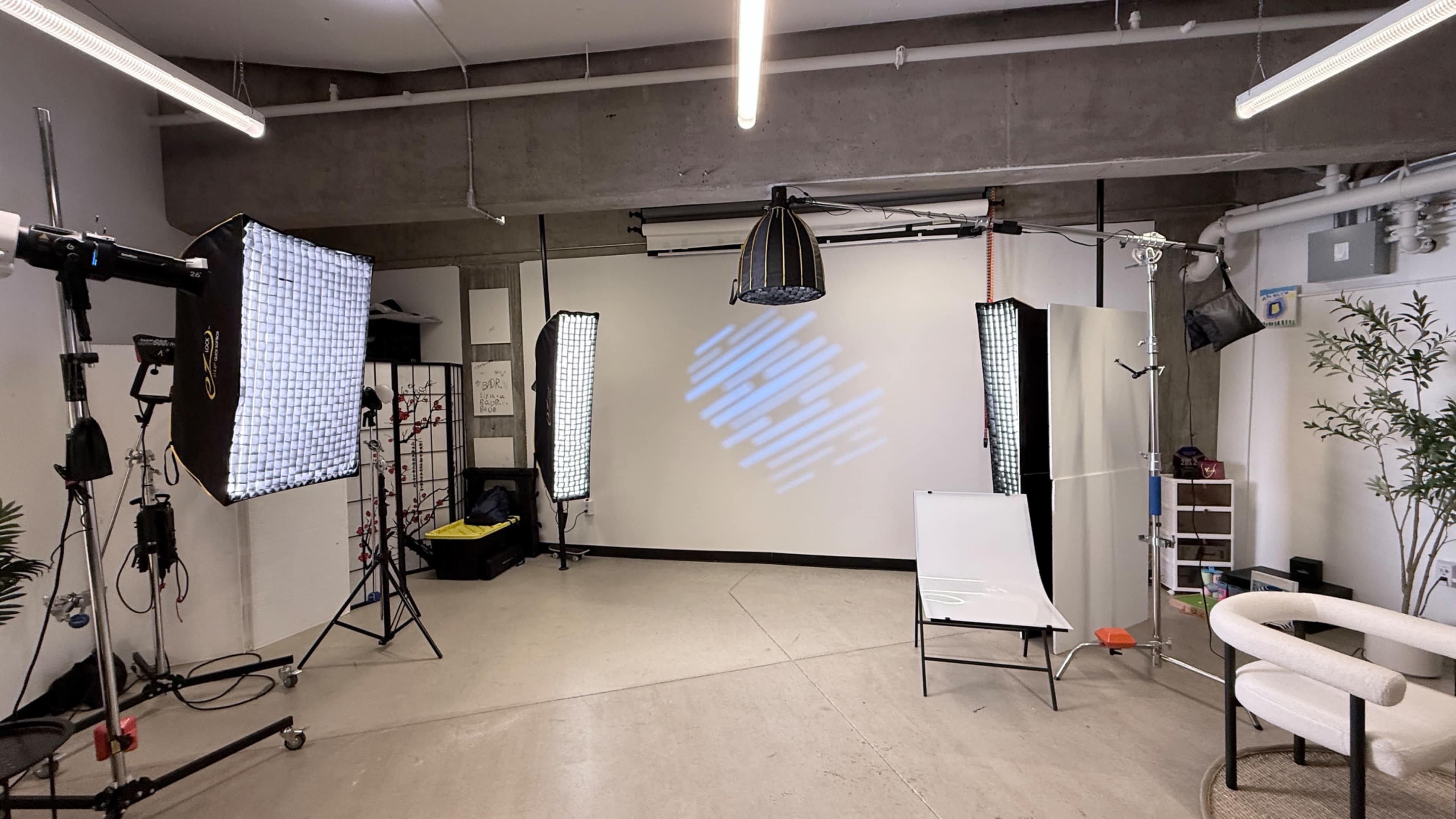 The image shows a well-equipped photography studio featuring multiple light stands, a backdrop, a white chair, and an abstract light projection on the wall.