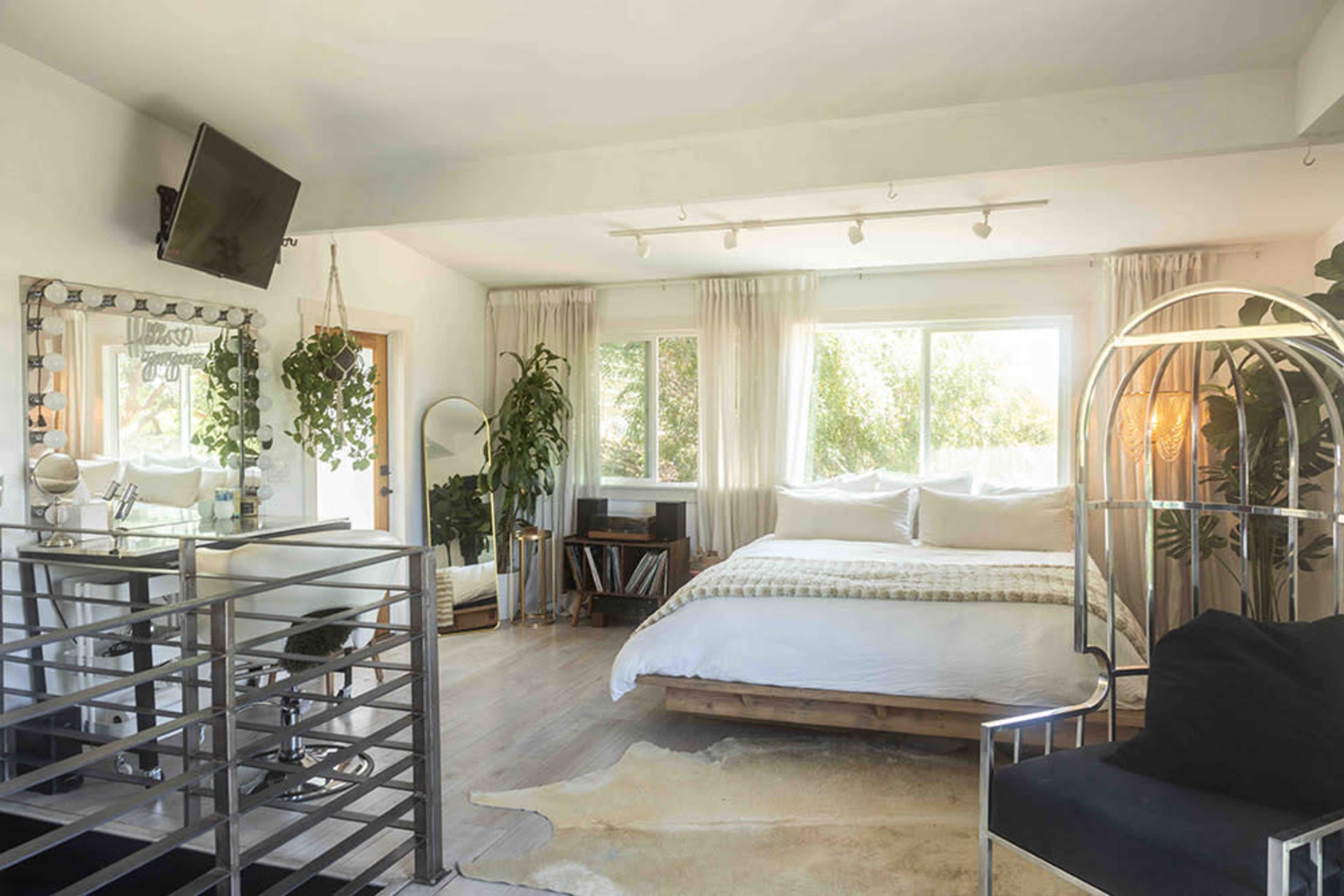 The image shows a well-lit bedroom featuring a large bed, a seating area with a chair, a mirror, and several indoor plants.