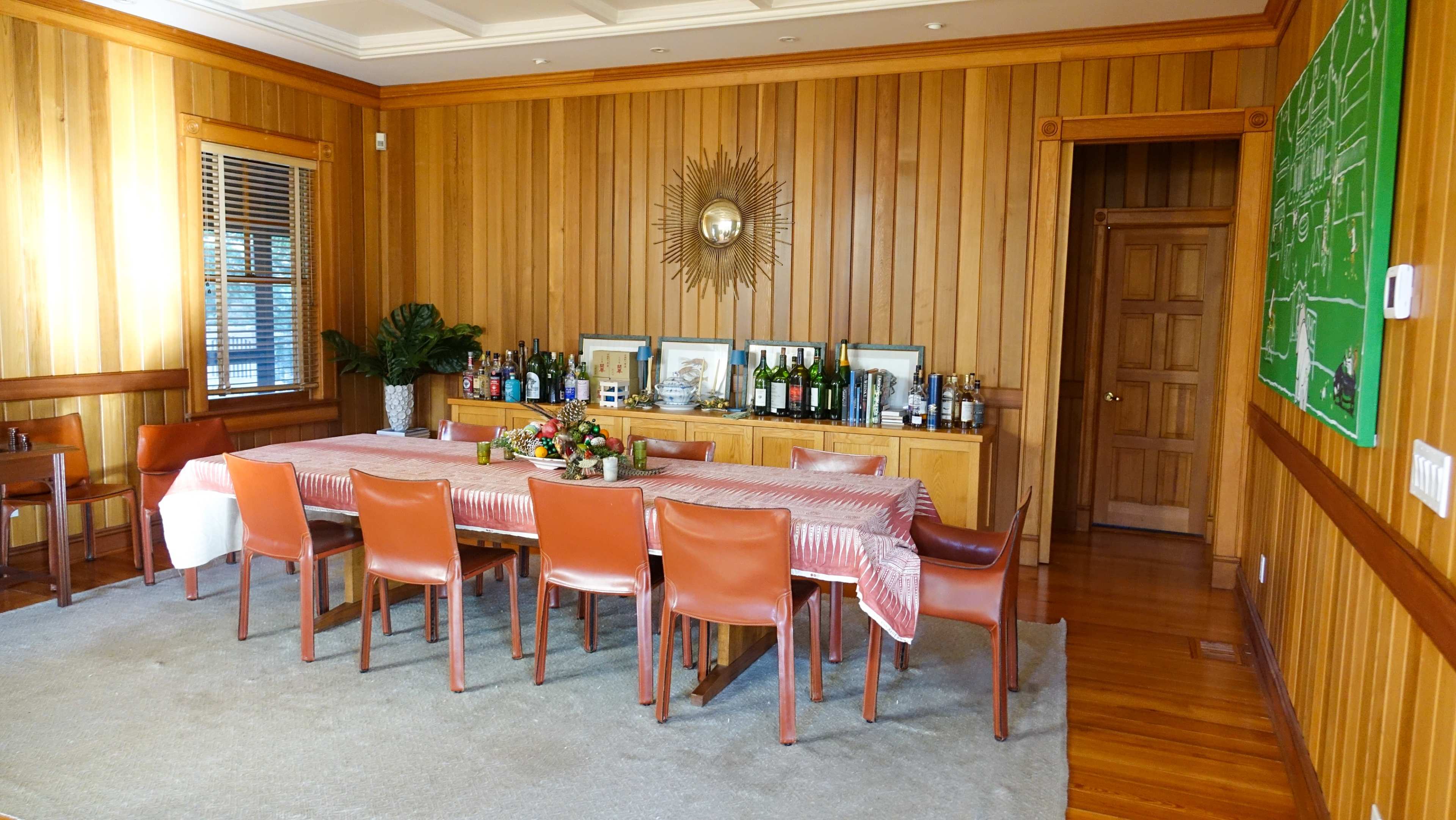 A wooden-paneled dining room with a long table set for a meal and a sideboard displaying various bottles and framed pictures.