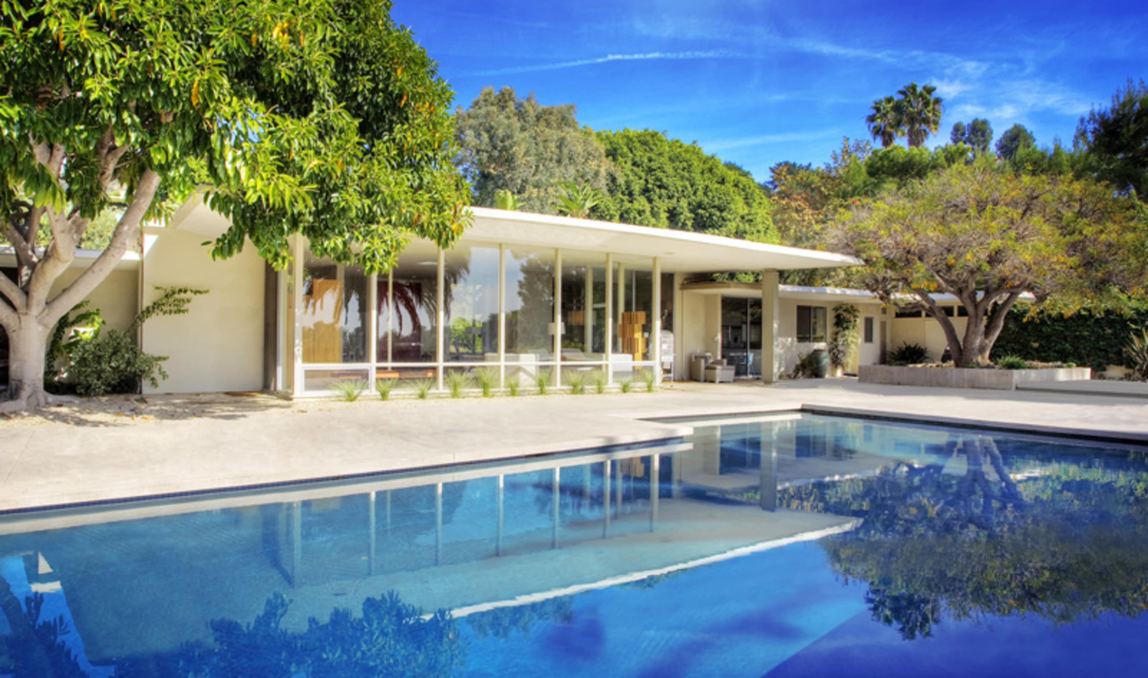 A modern house with large glass windows surrounded by trees and a reflective swimming pool in the foreground.