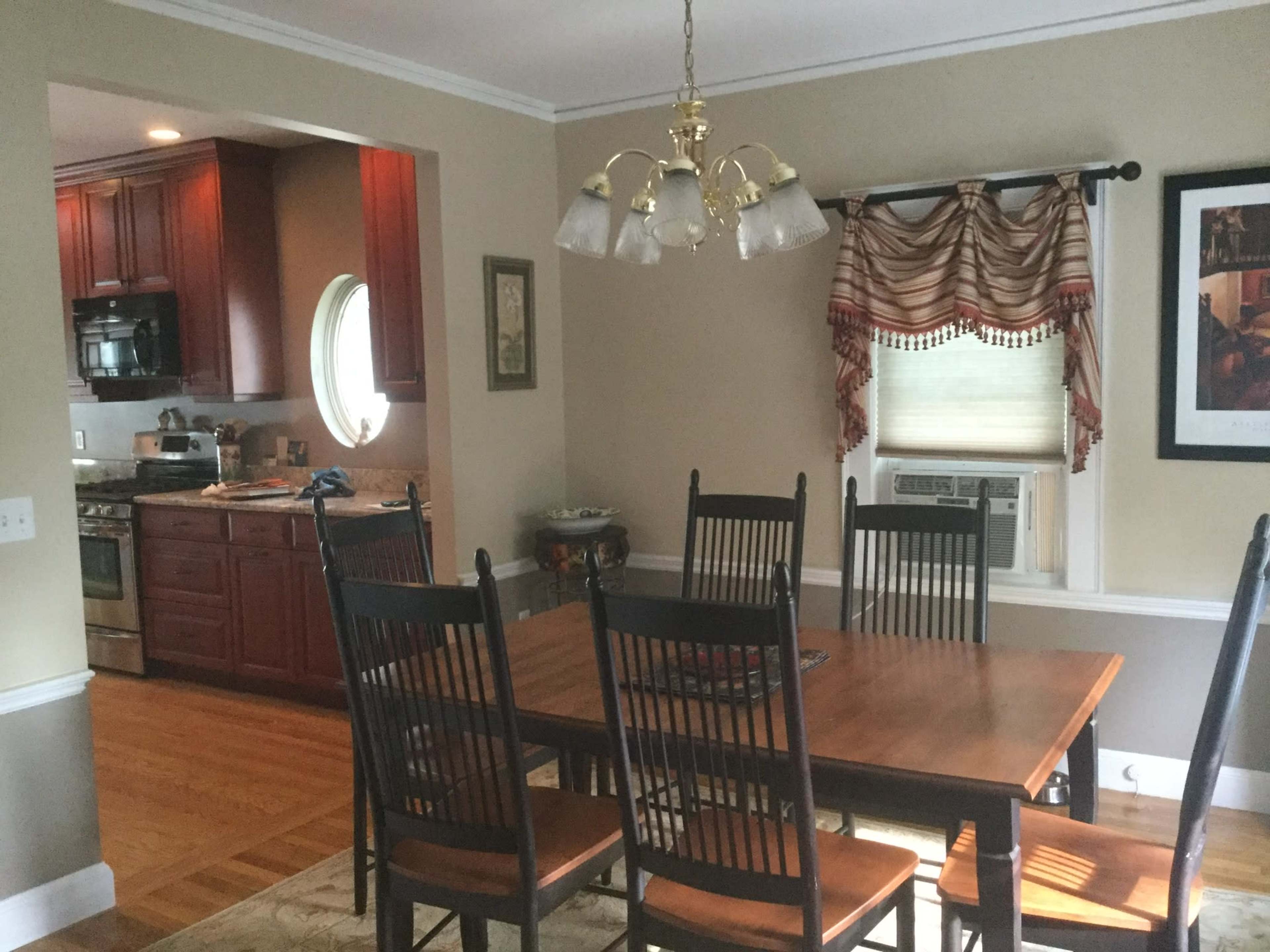 A dining room features a wooden table surrounded by black chairs, with a view of a kitchen in the background.