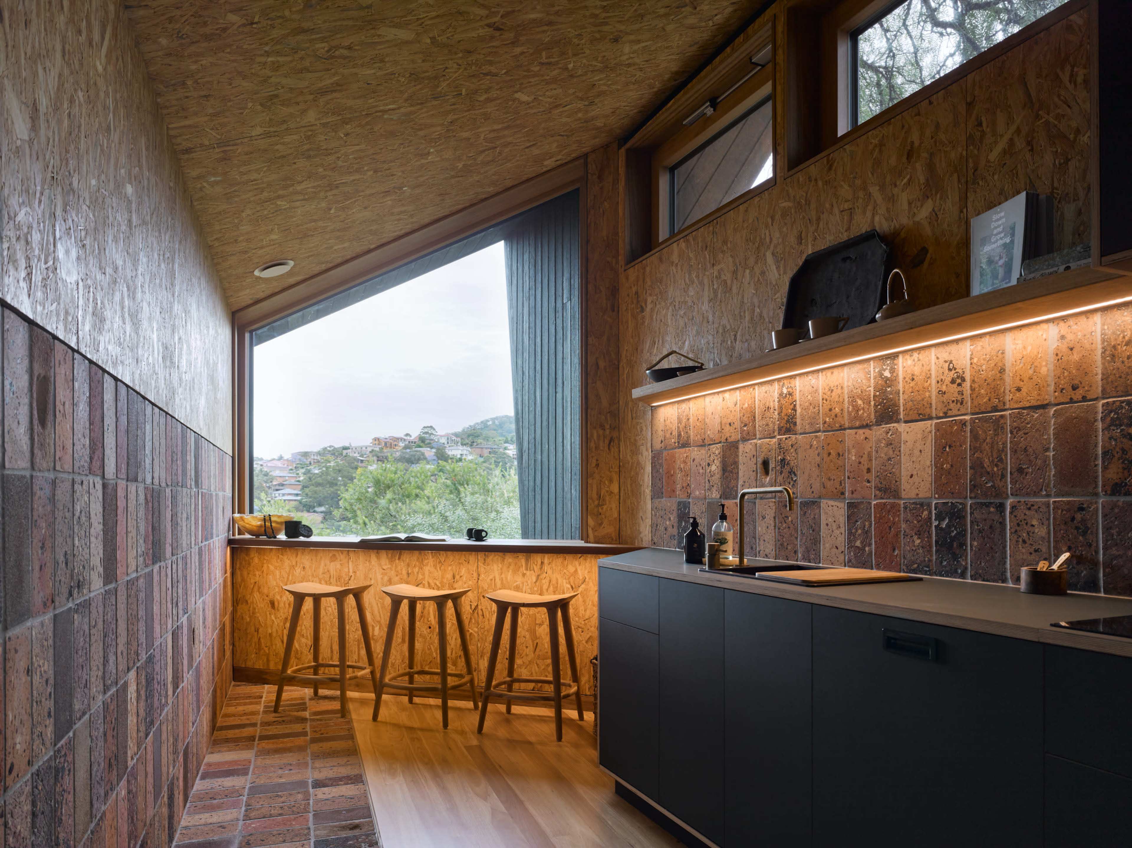 The image shows a modern kitchen with a sleek design, featuring a counter, wooden stools, and large windows that overlook a scenic view.