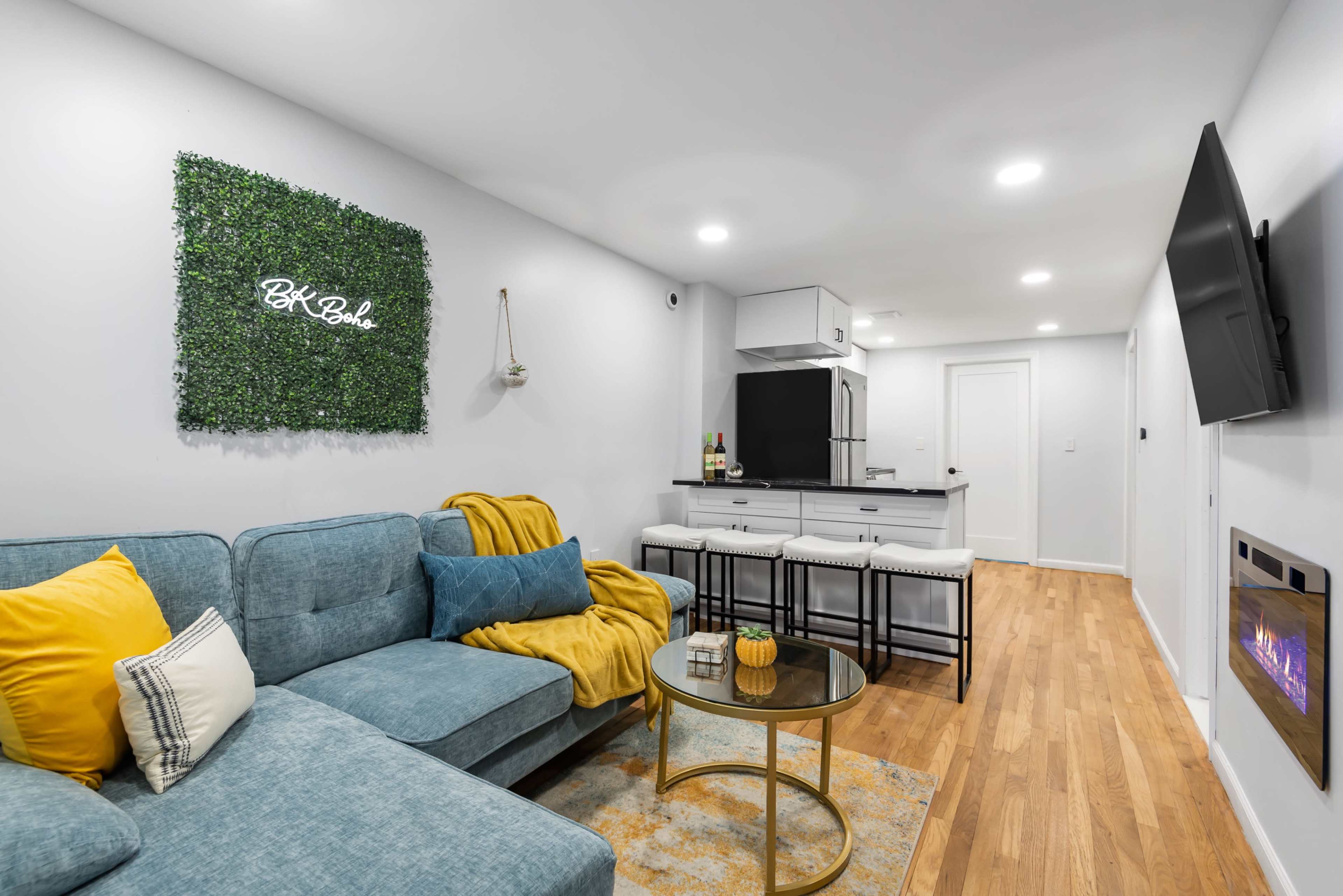 A modern living area with a light gray wall, a teal sectional sofa, a round glass coffee table, and a kitchenette in the background, featuring a bar area with stools.