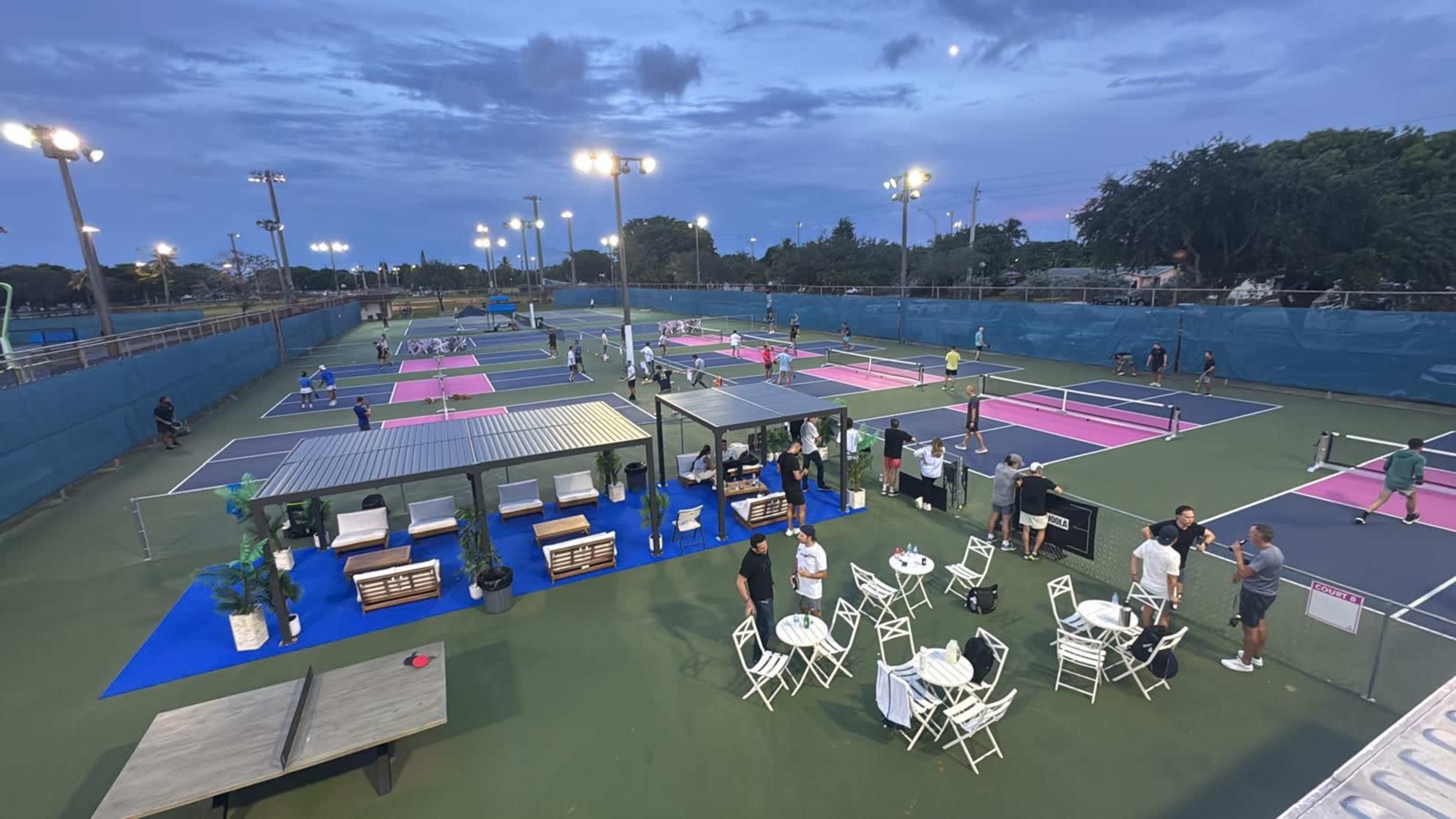 The image shows multiple tennis courts with players engaged in matches, surrounded by seating areas and spectators against a twilight sky.