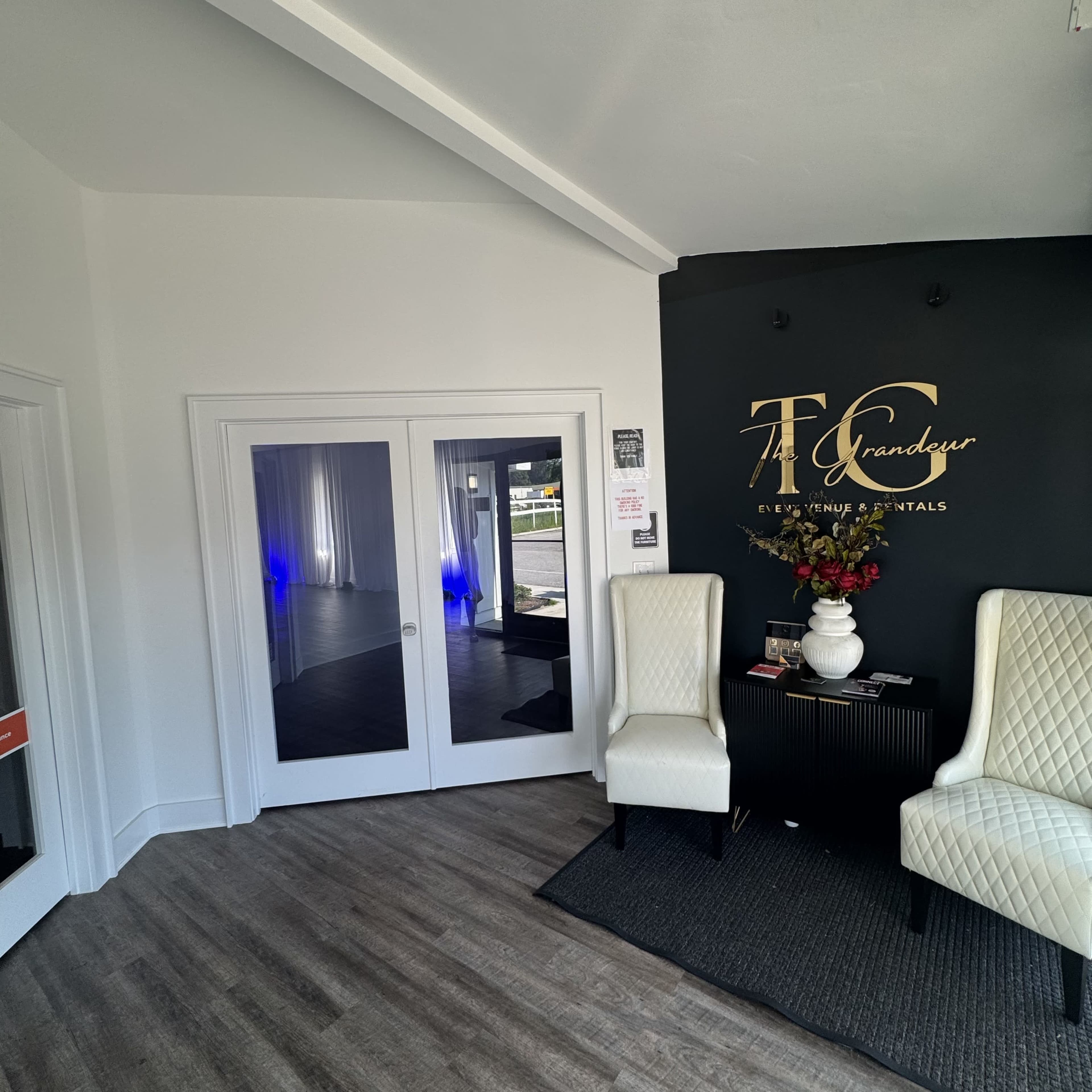 The image shows an interior space featuring two white chairs next to a dark wall with the text "The Grand" and a doorway leading to another room.