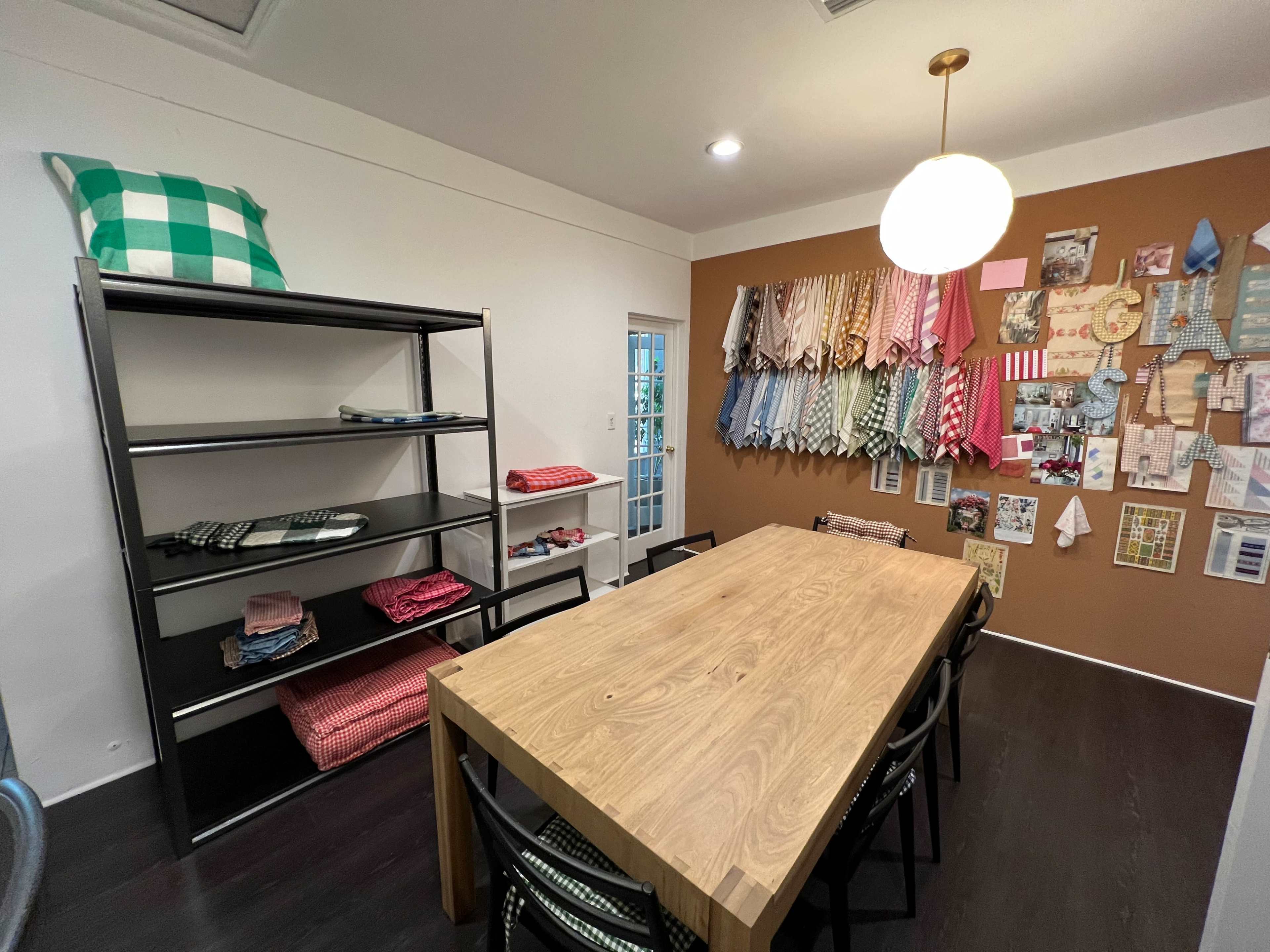 The image shows a bright room with a wooden table surrounded by chairs, a metal shelf on one side, and a wall displaying various fabric swatches and organized notes.