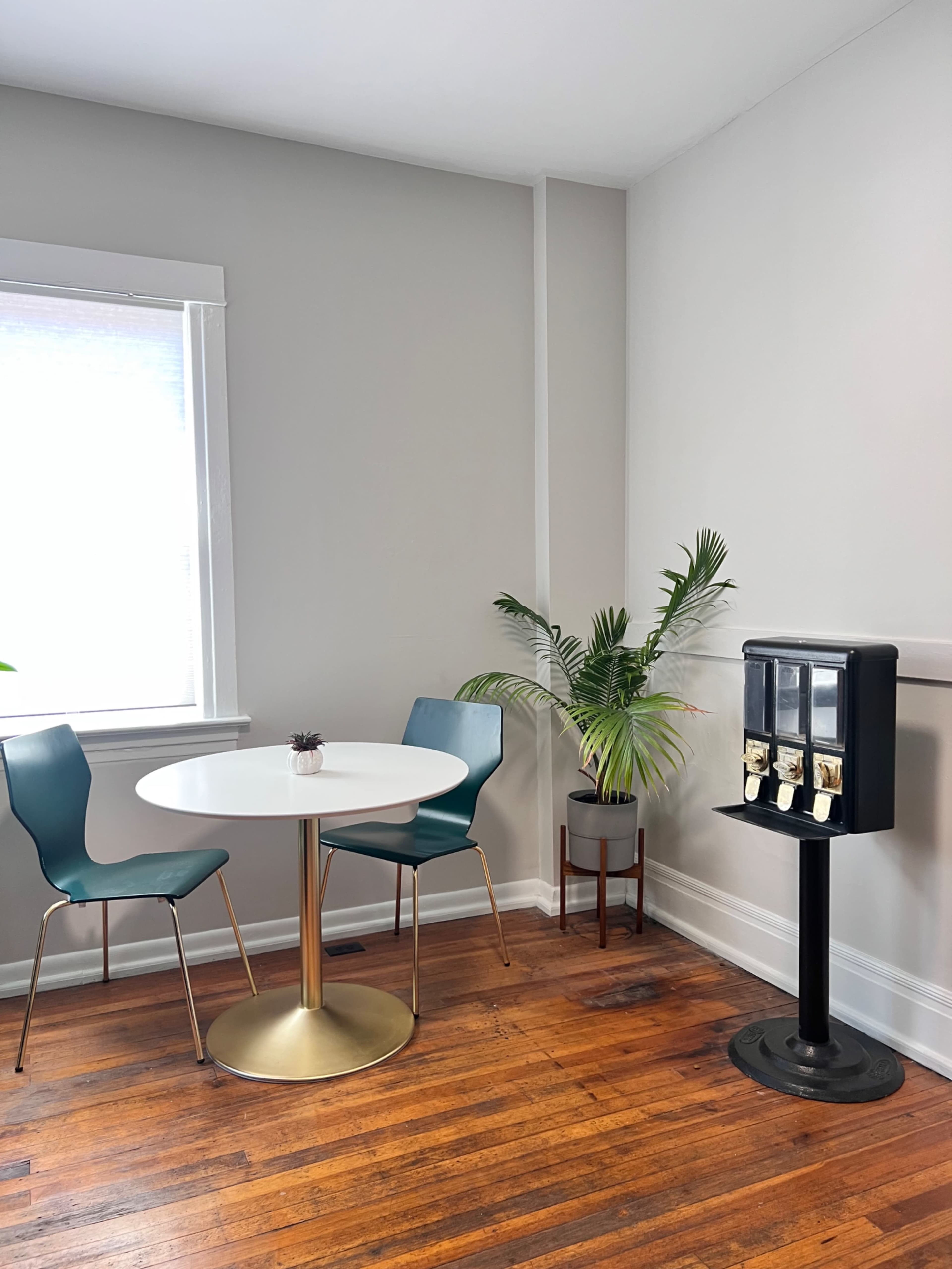 A small dining area features a round white table with three chairs, a potted plant, and a vending machine against a wall with light-colored paint.