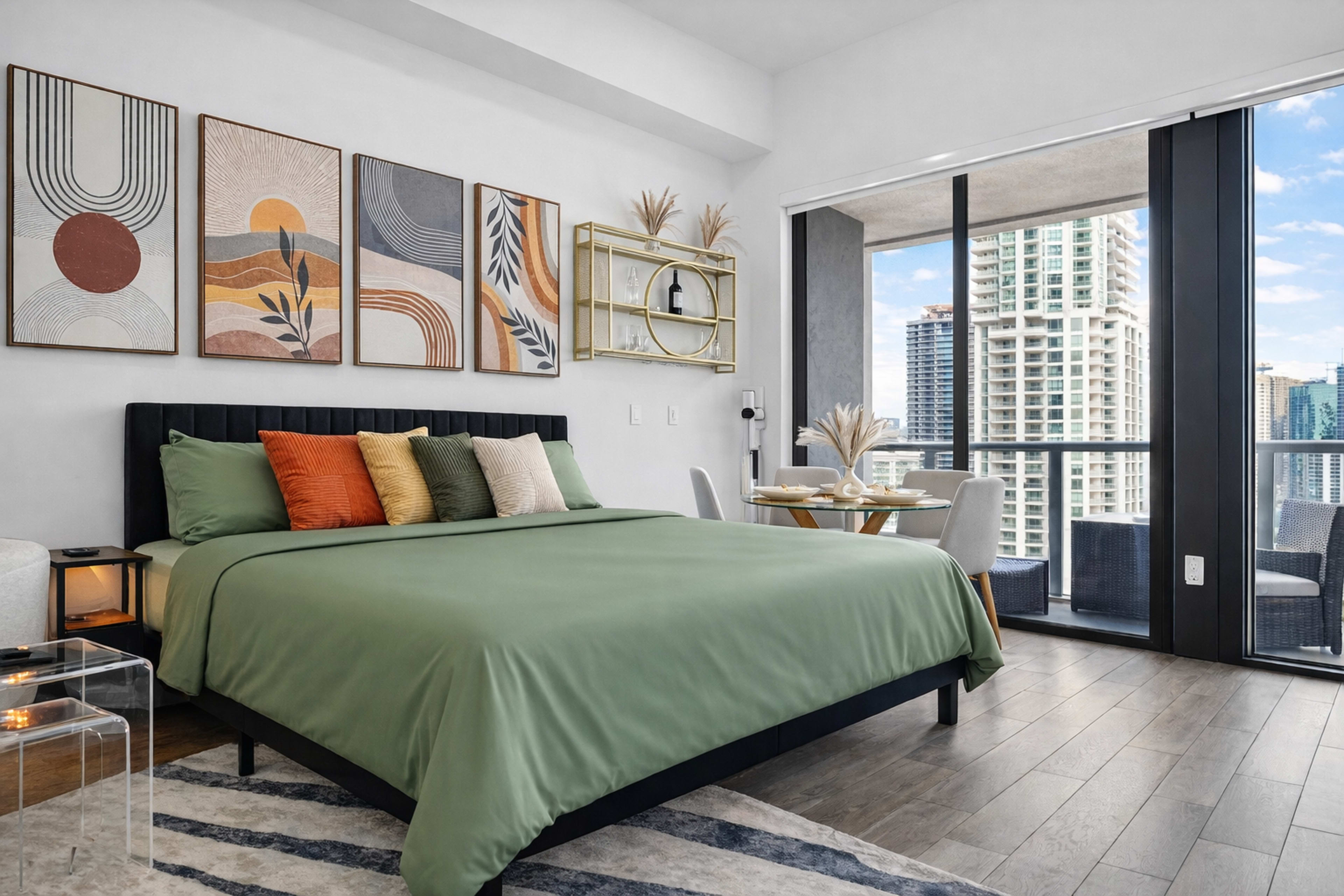 The image shows a modern bedroom with a green bedspread, decorative pillows, abstract wall art, and a balcony view featuring city skyscrapers.