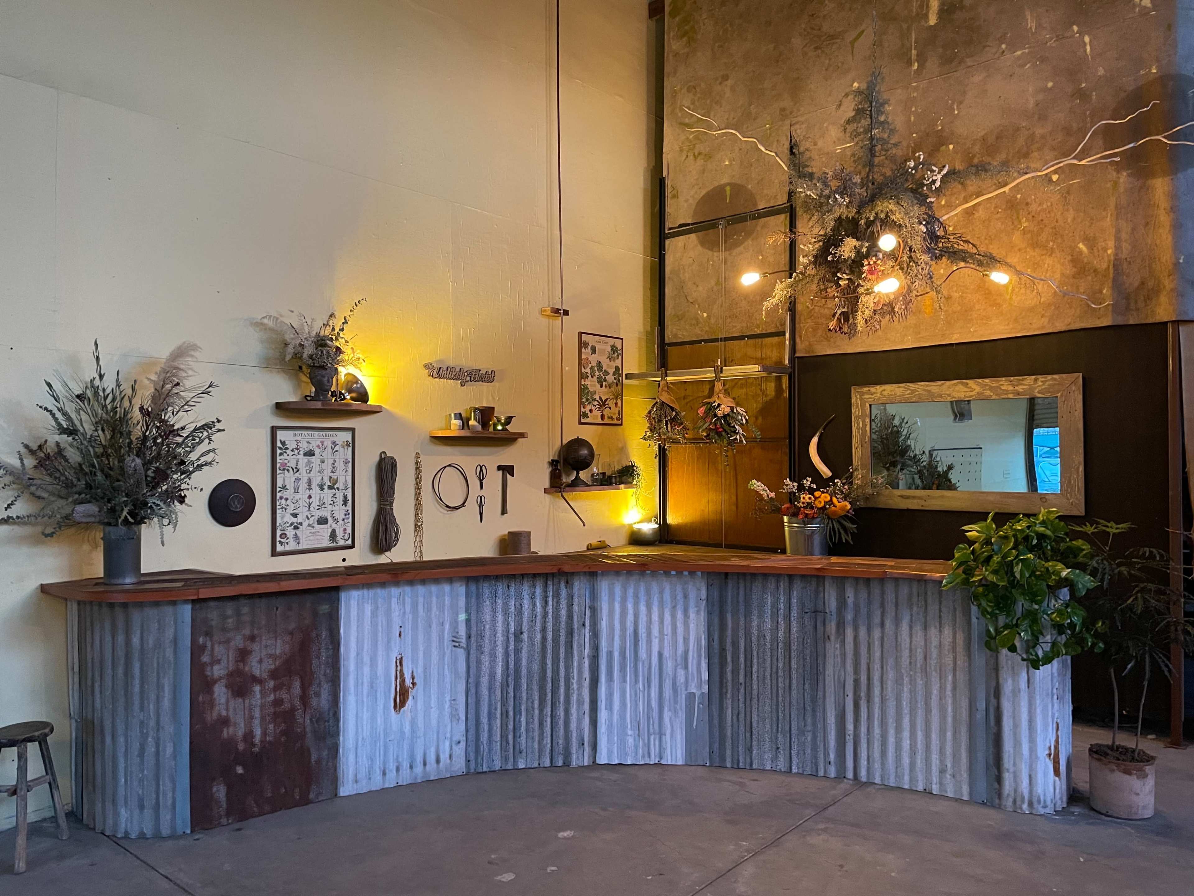 The image shows a curved reception desk made of corrugated metal and wood, surrounded by decorative plants and wall-mounted art in a well-lit, minimalist space.