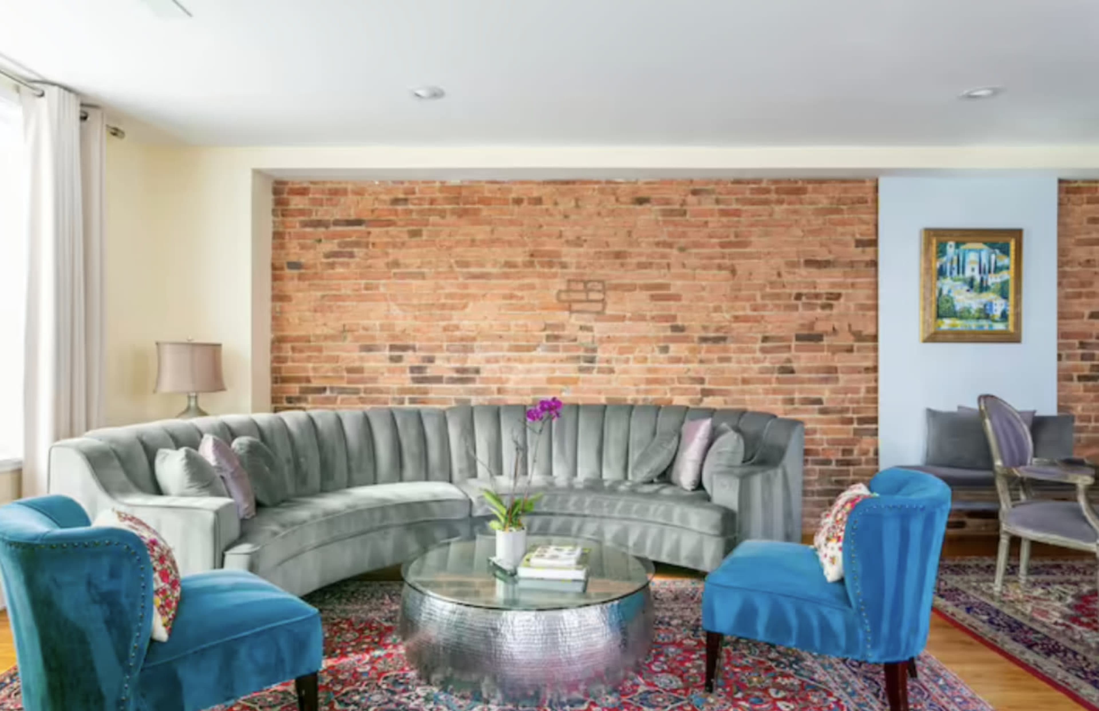 A living room featuring a curved gray sofa, two blue chairs, a circular glass coffee table, and an exposed brick wall.